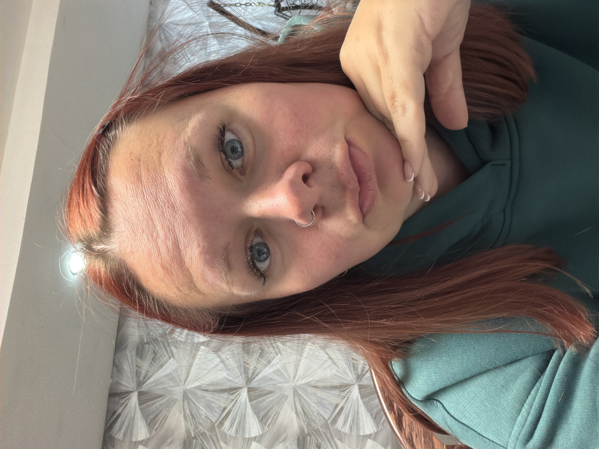 This media file is a close-up selfie of a young adult female, positioned with her head tilted, gazing directly into the camera. She has reddish-brown hair and light blue eyes. Her expression features a slight pout, and she wears a silver septum piercing in her nose. She is dressed in a teal or dark green hooded top. Her left hand, displaying manicured nails with white French tips, rests gently against her jawline and cheek.
The background suggests an indoor setting. To the left side of the frame, a plain, light-colored wall or surface is visible, with a bright, circular light source creating a strong reflection near her forehead. To the right, a textured wall featuring a geometric pattern of repeating starburst or fan-like shapes in white or light grey provides a decorative backdrop. The lighting is bright and evenly illuminates her face. The image depicts a casual, posed moment, likely taken indoors in a private residence in Chaikovsky, Russia. There is no visible text.