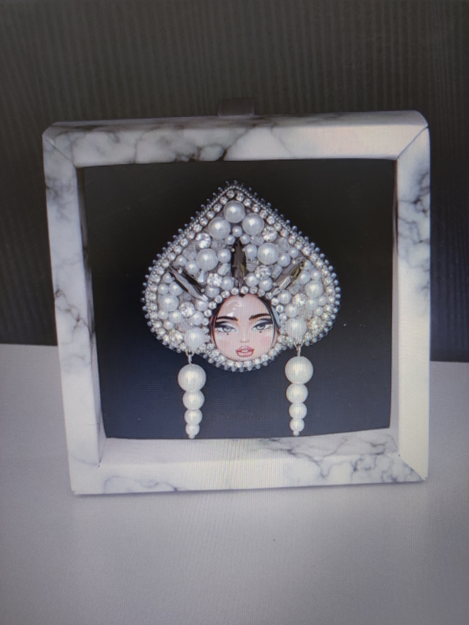A framed piece of art is displayed against a dark background. The art depicts a portrait of a woman wearing an ornate headdress adorned with pearls and diamonds. The headdress is large and heart-shaped, with dangling pearl earrings. The woman's face is rendered in a stylized manner, with prominent eyes and lips. The frame has a marble-like texture. The background appears to be a wall with horizontal lines. The overall scene is dimly lit, suggesting it might be an indoor setting. No people are present in the immediate vicinity of the artwork. The location in Pospelikha, Russia, is not visually evident from the image.