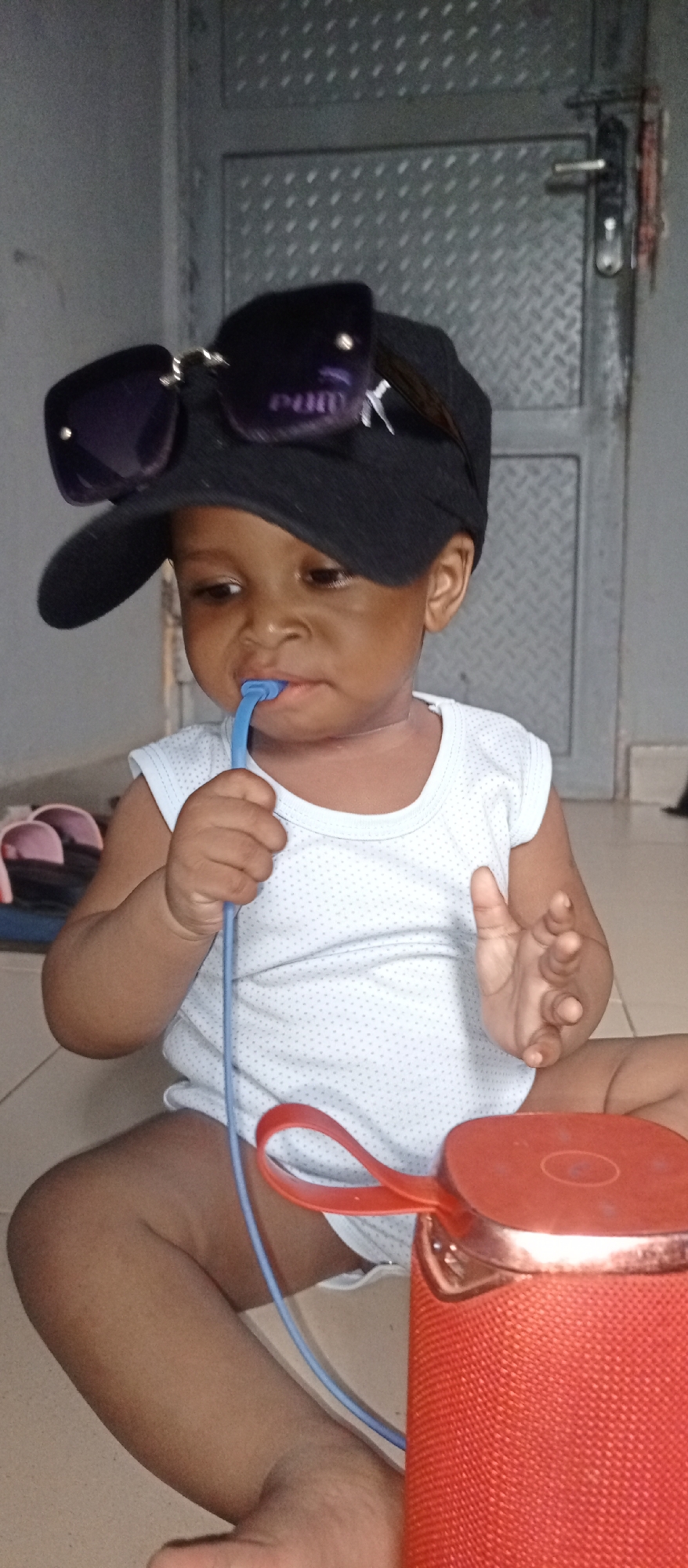 A young child sits on a tiled floor. The child is wearing a white sleeveless shirt, a black baseball cap with sunglasses perched on top. The child is holding a blue cord in his hand and putting the end of it in his mouth. Beside the child is a red portable speaker. In the background, there is a metal door.