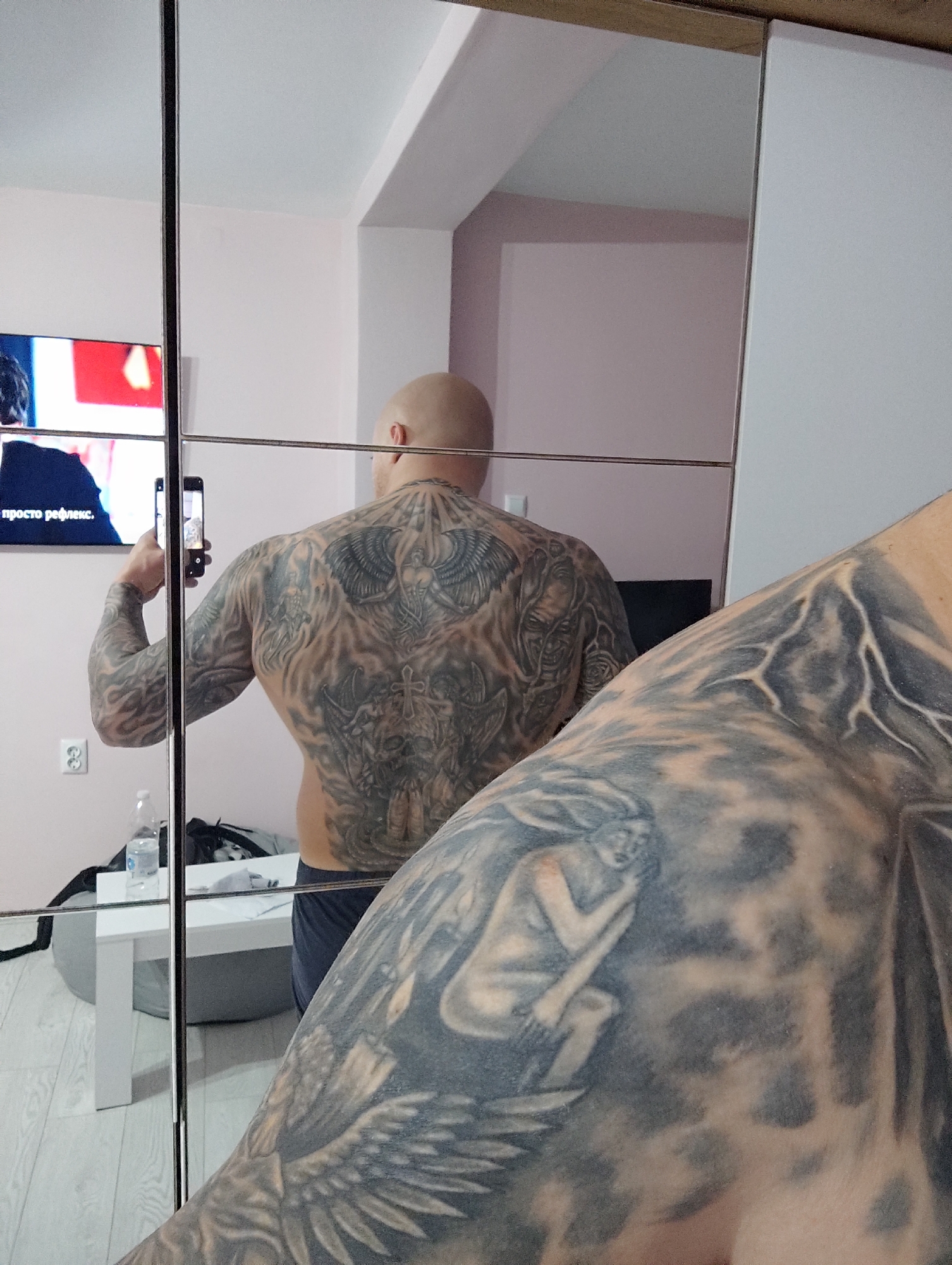 A bald man is taking a photo of his back tattoo in a mirror. The tattoo features images of winged figures and skulls. A television is visible in the background showing writing in a Cyrillic alphabet, and a white table with a water bottle sits in the lower left corner. The location is likely an interior space in Burgas, Bulgaria. A tattoo of a woman and an eagle covers the foreground.