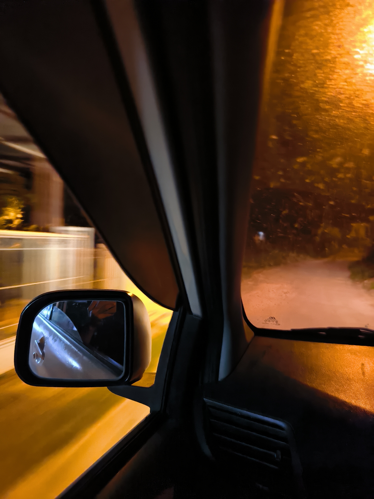 The media file shows a view from inside a car at night, looking out the passenger side window and at the side mirror. The scene outside is blurred due to motion, suggesting the car is moving. Streetlights cast a warm, orange glow on the wet road and vegetation, indicating it might be raining or has recently rained.
In the side mirror, a reflection of the car's interior is visible, including what appears to be a person's arm and shoulder. The exterior of the car door is also reflected.
The overall mood is somewhat abstract and atmospheric due to the low light, motion blur, and reflections. The time of day is clearly night. The presence of streetlights and a road suggests an urban or suburban environment. The wetness on the road and possibly on the window implies damp weather conditions. There is no discernible text in the image. The location context provided is Lhokseumawe, Indonesia.