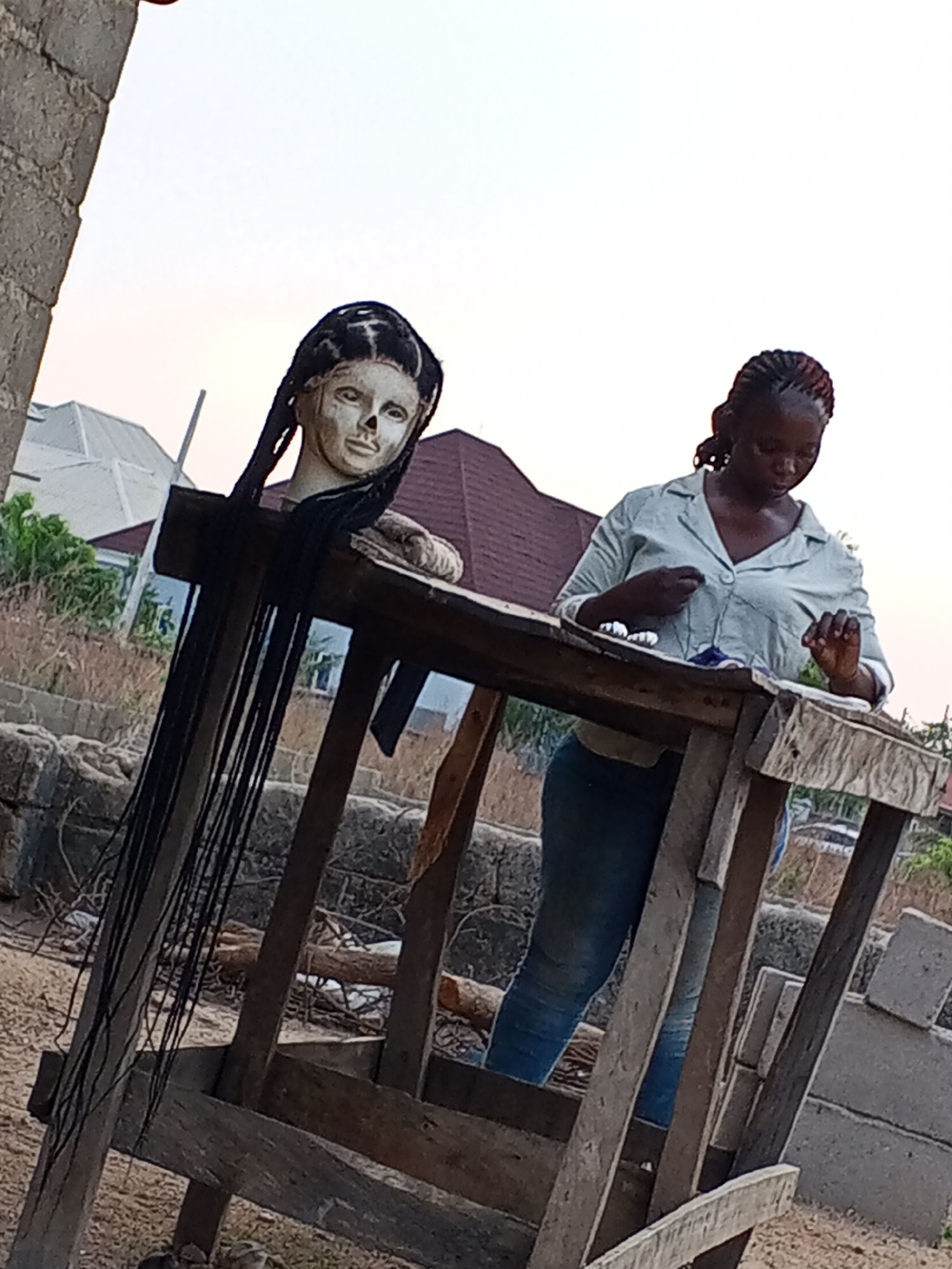 The image depicts a woman standing behind a makeshift wooden table in what appears to be an outdoor workspace in Adonkolo, Nigeria. She is wearing a light-colored, button-down shirt and blue jeans and seems to be engaged in a detailed handiwork task at the table. To her left, on the same table, there is a mannequin head with long, dark braided hair. The table itself is rudimentary, constructed from rough-hewn wooden planks and posts. The background shows a mix of residential buildings, including what appears to be a white house with a gray roof and some smaller structures. The environment is somewhat cluttered with debris, suggesting a working area that is not particularly well-maintained. The overall lighting suggests it is either early morning or late afternoon.