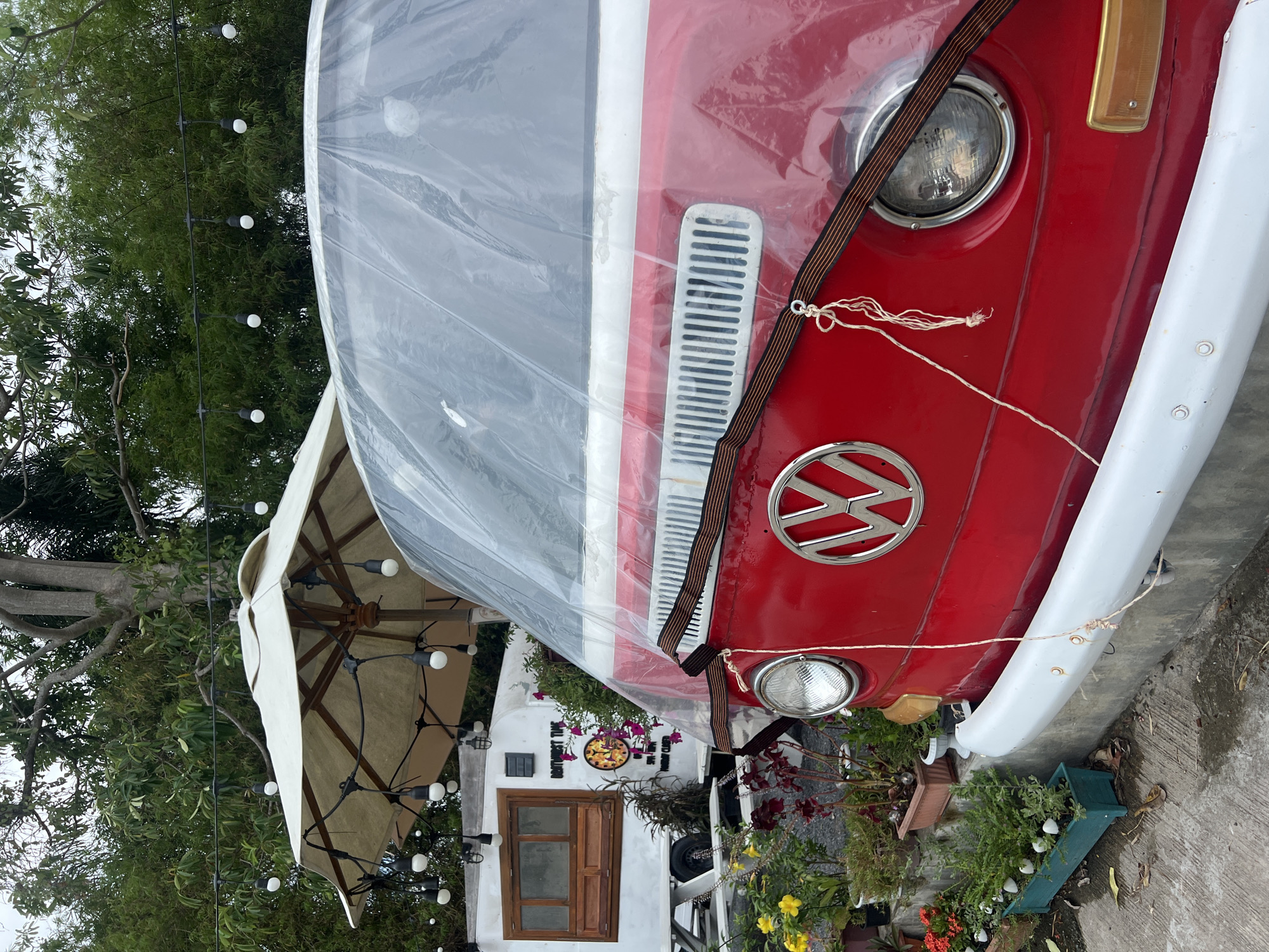 An outdoor scene features a red and white vintage Volkswagen Type 2 van, parked and partially covered by a clear plastic sheet. The van's front, including its circular headlights and the prominent "VW" logo, is visible. A thin rope is tied around the plastic cover, securing it. To the left of the van, a large beige outdoor umbrella is open, its inner structure with wooden spokes visible. Numerous string lights with spherical bulbs are strung across the scene, appearing both in the foreground and extending towards the background, creating a festive ambiance.
Behind the umbrella and to the left of the van, a white building or structure with a rustic wooden door and a small wooden-framed window can be seen. On the wall of this building, a circular clock with a floral pattern and Roman numerals is present. Text is written on the wall; above the clock, it reads "DON'T WORRY, TIME," and below the clock, partial text appears to say "LIVE YOUR DREAMS" and "DO WHAT YOU LOVE."
The surrounding area is lush with greenery, including tall trees with dense foliage in the background and various potted plants and flowerbeds in the foreground. Some pots contain green leafy plants, while others display red and yellow flowers. The ground beneath the van and plants appears to be concrete or paved. The lighting suggests an overcast daytime setting, with no harsh shadows visible. The overall impression is that of a charming outdoor cafe or garden display area.