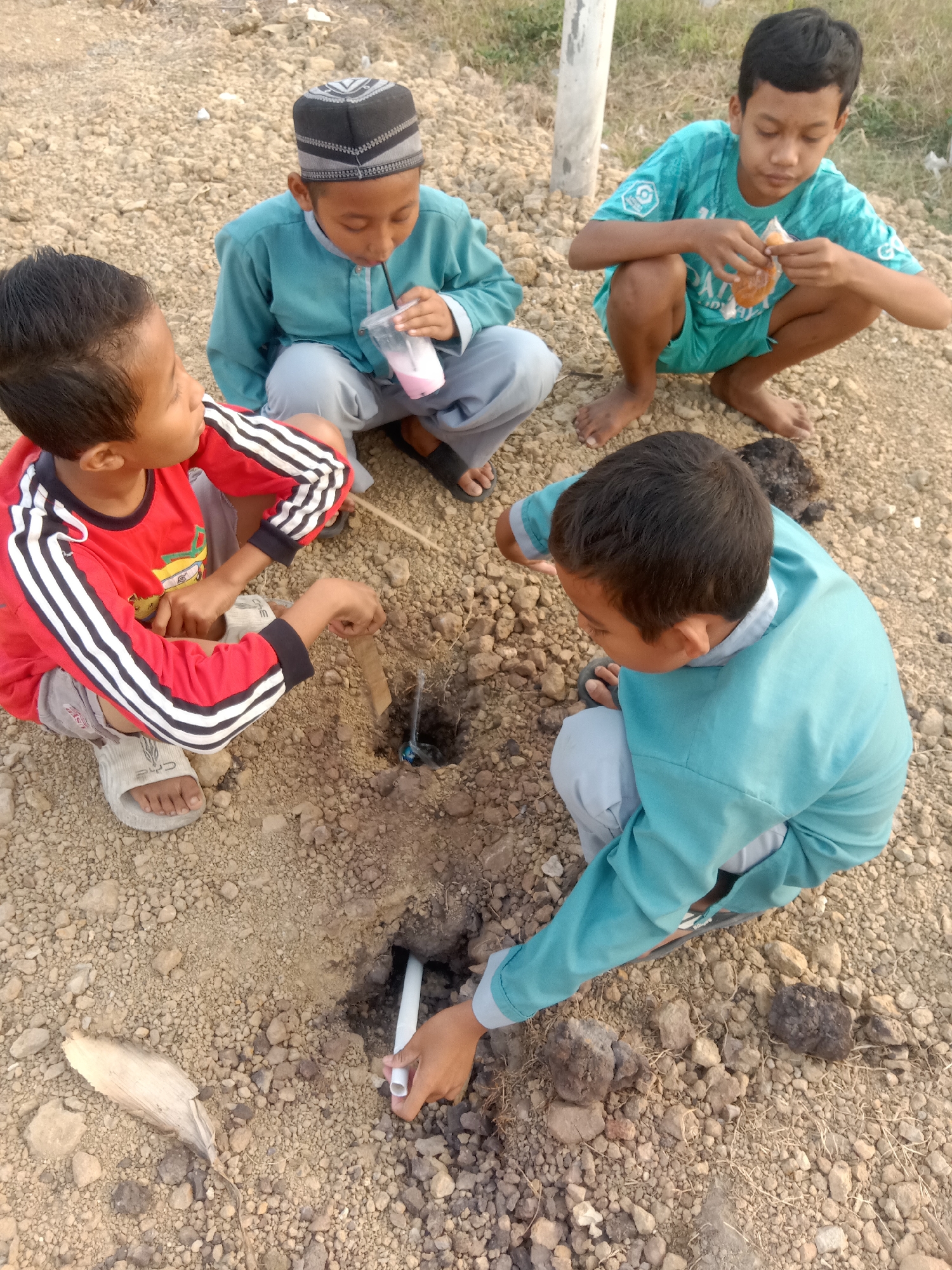 Four young boys are outdoors, crouched on a dirt and gravel ground. One boy, wearing a red shirt with black and white stripes and sandals, is focused on a hole in the ground, holding a piece of brown paper and a small object. Another boy, in a light blue long-sleeved shirt and pants, is also looking at the hole and holding a white pipe. To his left, a boy in a similar light blue outfit and a black and gray kufi is drinking a pink beverage through a straw from a plastic cup. The fourth boy, in a teal t-shirt and shorts, is crouched nearby and appears to be eating something. The environment is dry and appears to be an undeveloped area, possibly in Stiaji, Indonesia, given the presence of dry vegetation and loose stones.