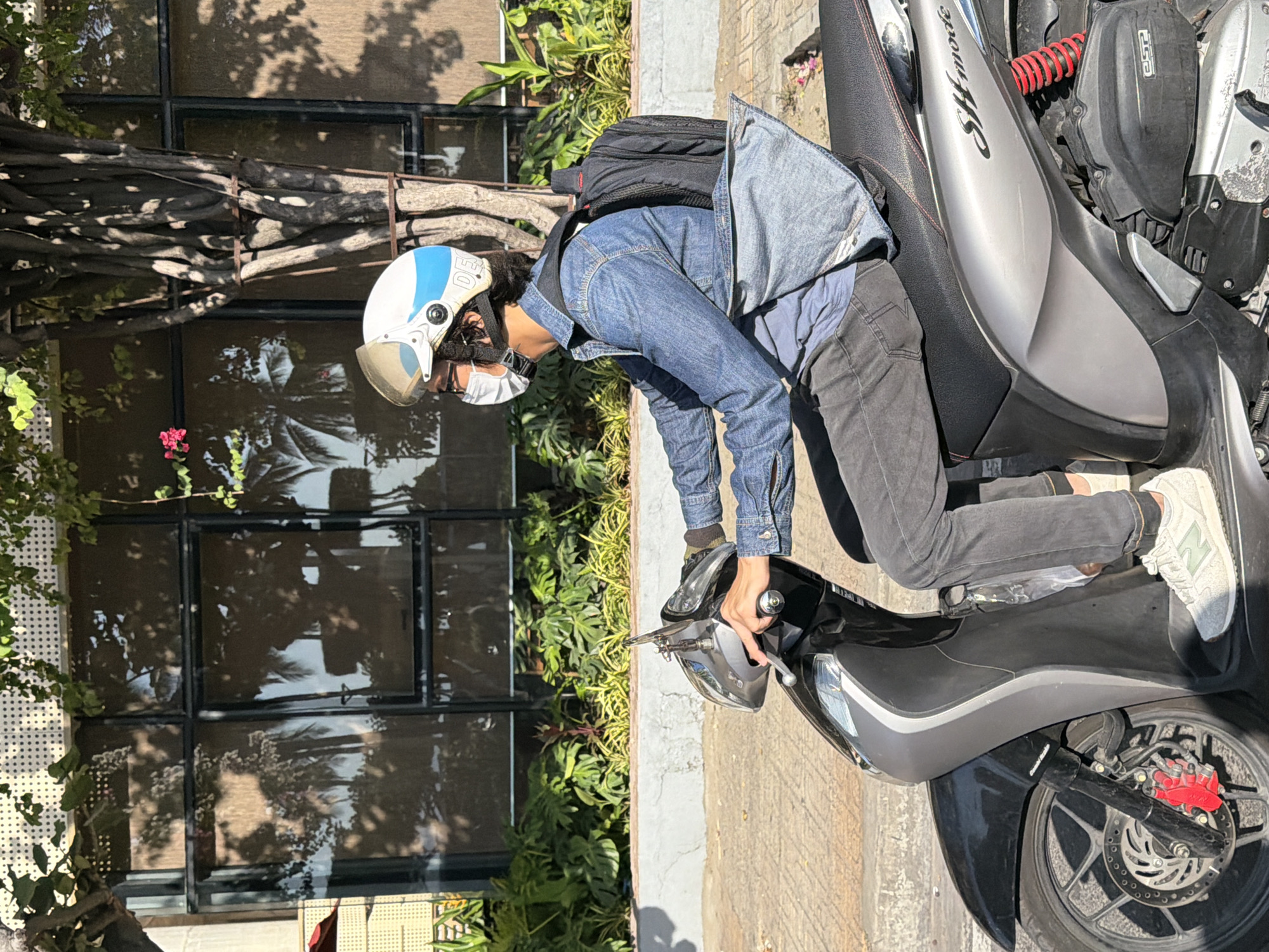 A person wearing a denim jacket, dark pants, a white face mask, and a white and blue helmet is riding a silver and black scooter. The person is positioned in the right side profile, with their right hand on the handlebar. A black backpack is visible on their back. The scooter has the "SH mode" branding on its side. The scene appears to be on a street in Nha Trang, Vietnam, suggested by the presence of palm tree reflections in the building windows and the general style of the architecture and vegetation. The background features a building with large windows reflecting palm trees and foliage, as well as a tree with textured bark. The ground is paved, and there is a grey curb and planted greenery. The lighting indicates it is daytime, with strong shadows cast by the sun.