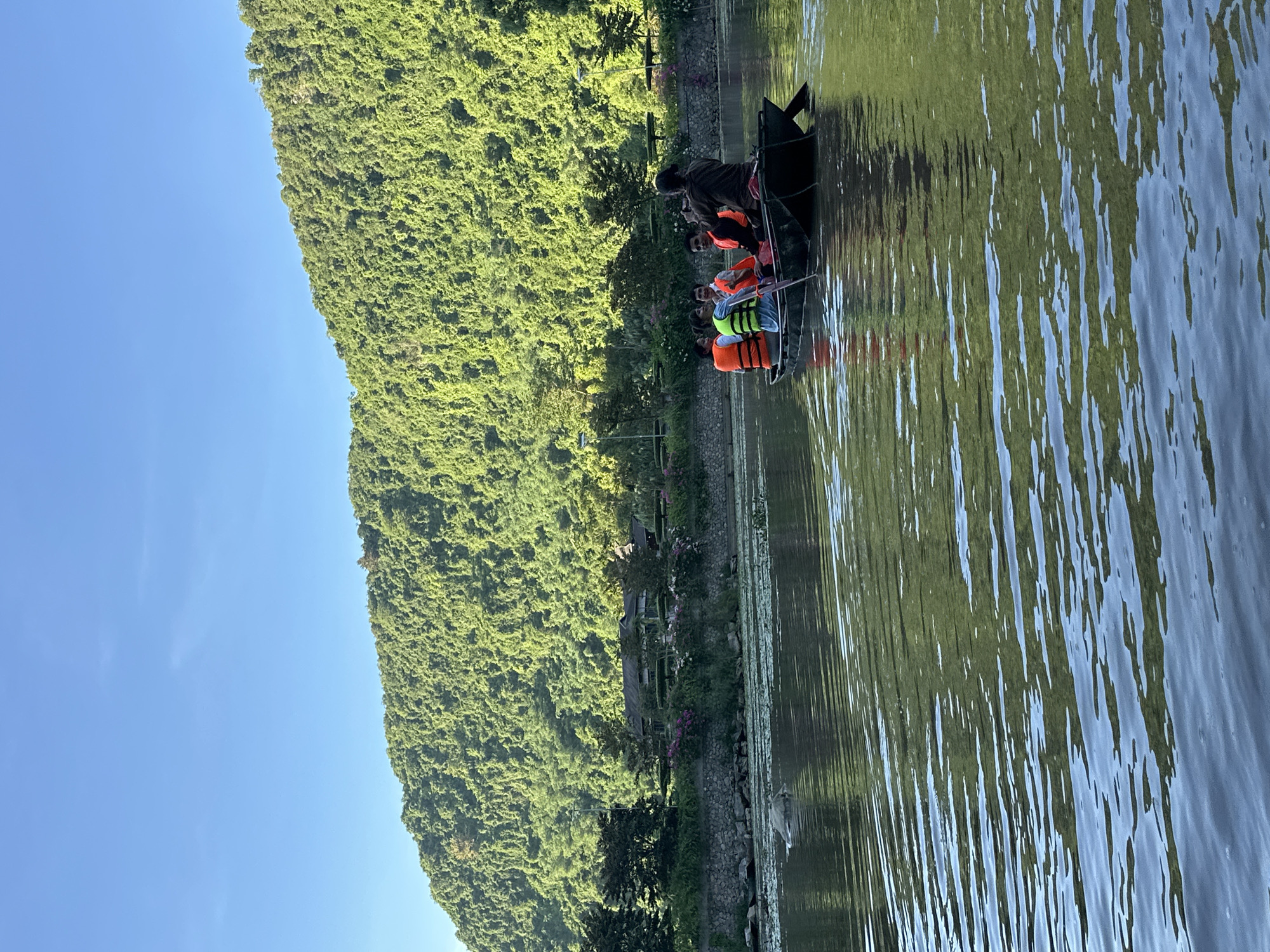 In the city of Yen Son Ward, Vietnam, a small boat navigates a calm body of water. The boat, made of dark material, carries five individuals, all wearing life vests in vibrant orange and green. One person at the rear of the boat appears to be steering with an oar. The water's surface reflects the surrounding lush green hills that rise majestically behind a treeline and a stone wall lining the shore. A building with a dark roof is visible nestled amongst the trees on the hillside. The sky above is a clear, bright blue, suggesting fair weather. Lily pads are scattered across the water in the foreground.