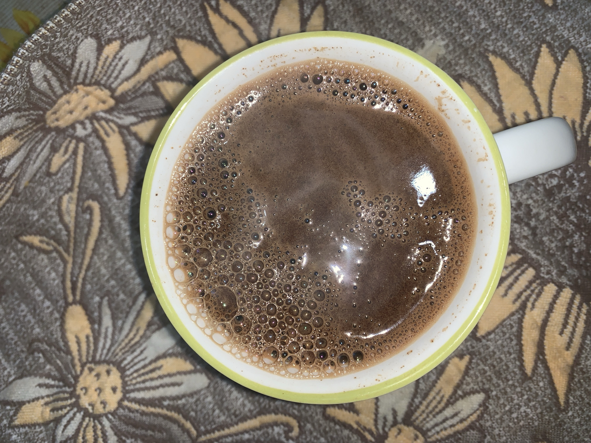 A close-up, overhead shot shows a mug of hot chocolate with a foamy surface. The mug has a white interior and a lime-green rim. The mug rests on a patterned fabric, which appears to be a tablecloth or placemat, featuring stylized yellow and brown flowers. The fabric has a dark brown background. The hot chocolate is a rich brown color with a multitude of small bubbles on its surface, some reflecting light. The image is focused on the mug and its contents, with the patterned fabric slightly blurred in the background. The scene is indoors, likely on a table.
