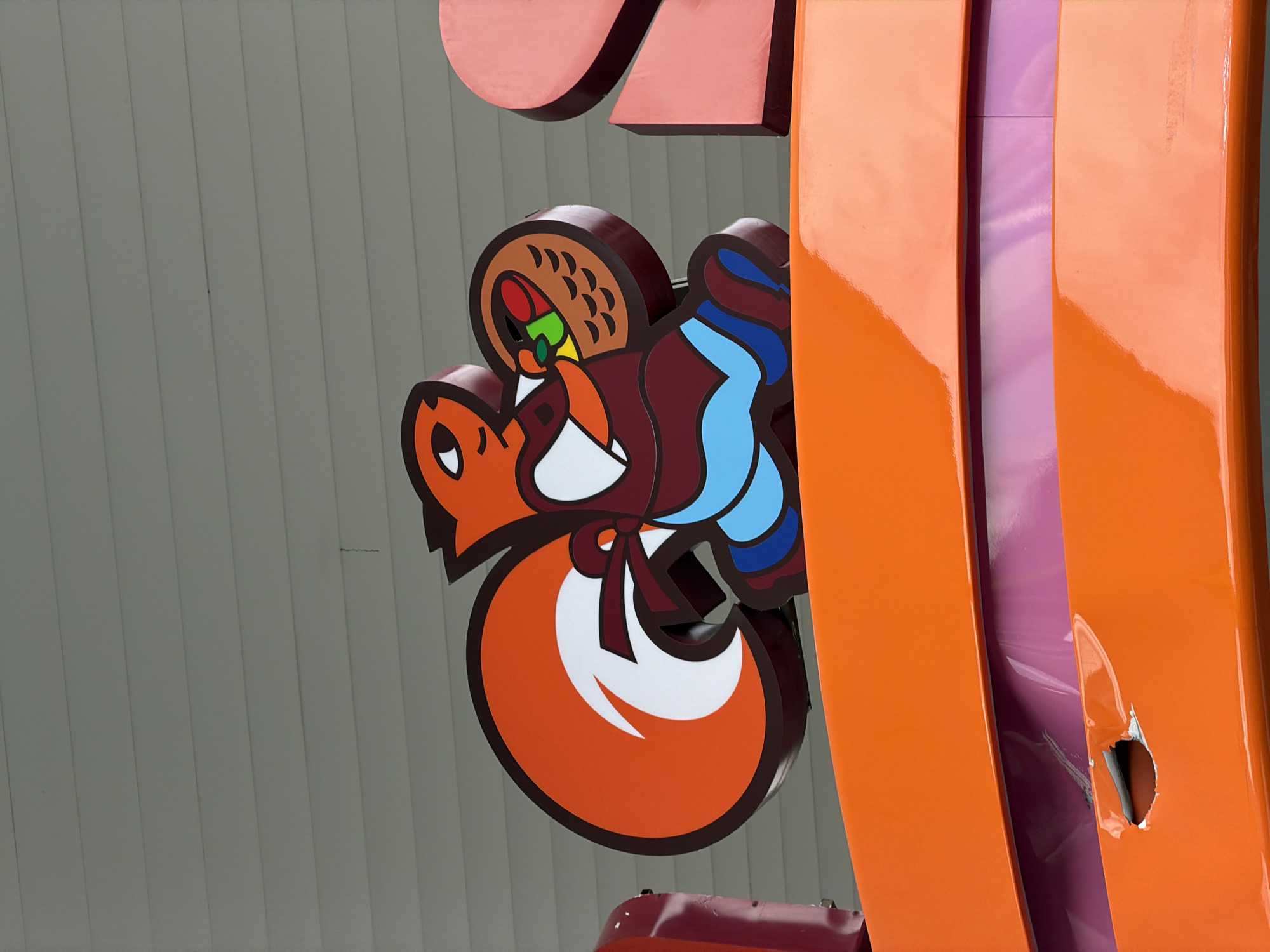 The image shows a portion of the "Taco Loco" sign in Tbilisi, Georgia. The sign includes a cartoon fox holding a taco, next to an orange element with a hole in it, exposing a purple layer underneath. The background is a light gray wall with vertical lines.