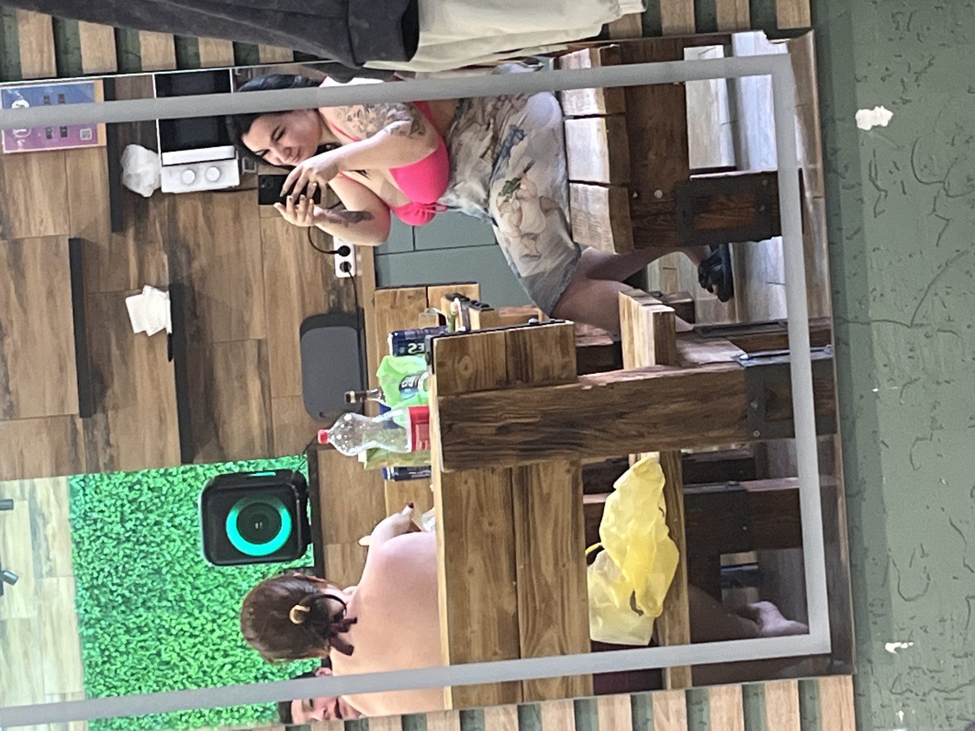 The image is a reflection in a mirror, depicting two women in what appears to be an indoor setting. One woman is wearing a pink bikini top and a floral print wrap, holding a mobile phone as if taking a picture of the mirror reflection. The second woman is partially visible, with her hair in a bun and appearing bare-chested.
The setting includes wooden furniture and shelves with various items such as water bottles, cleaning supplies, and a green square object with a circular green light. There is a wooden tiled wall behind the mirror. The information does not provide enough data to conclude the location in the city of Chaikovsky, Russia.