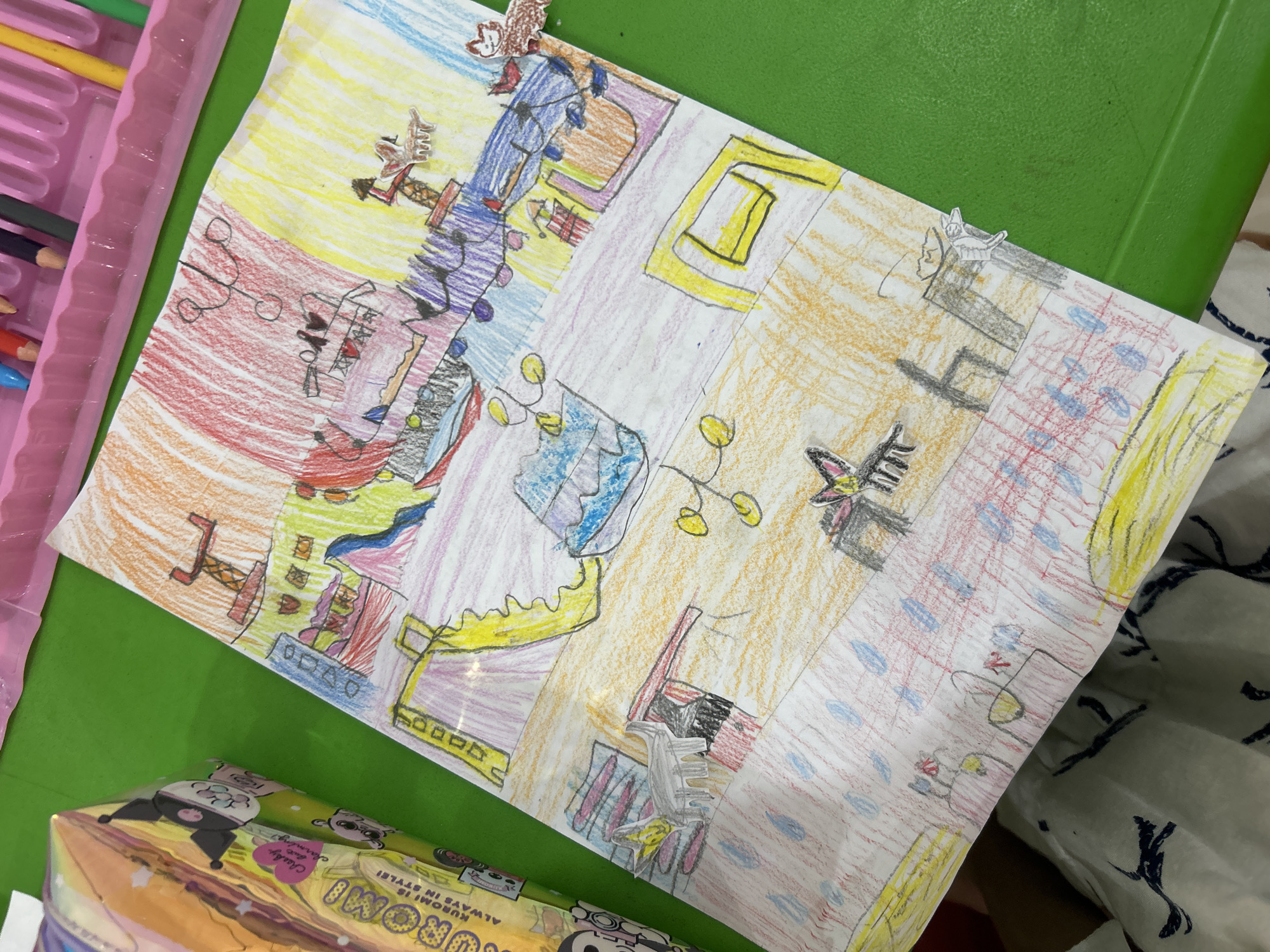 This is a photograph of a colorful, child-like drawing on a piece of white paper, resting on a green surface. The drawing depicts various abstract shapes and figures rendered in crayon, with different sections divided by horizontal lines. The top section of the drawing features red and orange hues, with what appears to be a structure resembling a tower. Below this, sections are colored in yellow, blue, and purple, with suggestions of furniture and decorative elements like lights. There are also small, cutout figures of animals or characters attached to the drawing in various locations. To the left of the drawing, a portion of a product packaging is visible, featuring a cartoon character with black ears and pink cheeks. The green surface could be a table or a desk. The drawing is not located in Kuta, Indonesia.