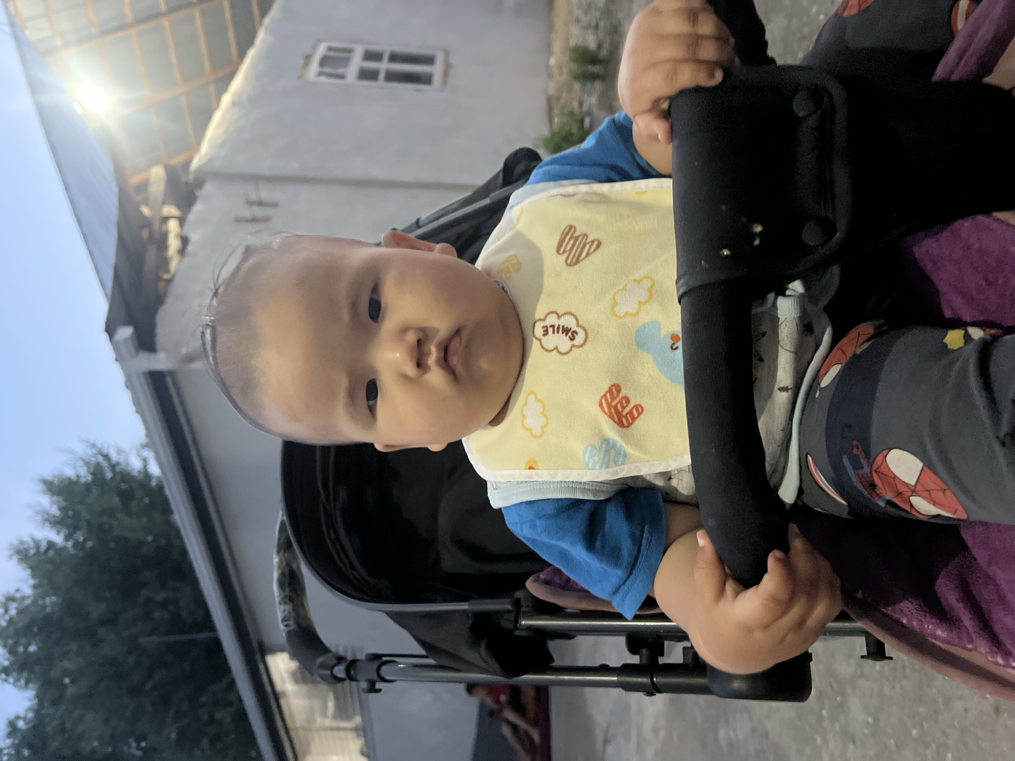 A baby is seated in a stroller, holding onto the stroller's handle. The baby is wearing a blue shirt and a yellow bib with cartoon cloud and heart designs. The background shows a building with a corrugated metal roof and windows. The scene appears to be outdoors, possibly in a residential area, given the building's structure. The time of day seems to be dusk or evening, as suggested by the lighting conditions. The overall mood is calm, with the baby looking directly at the camera. The image was taken in Kara-Dıykan, Kyrgyzstan. There is no visible text in the image.