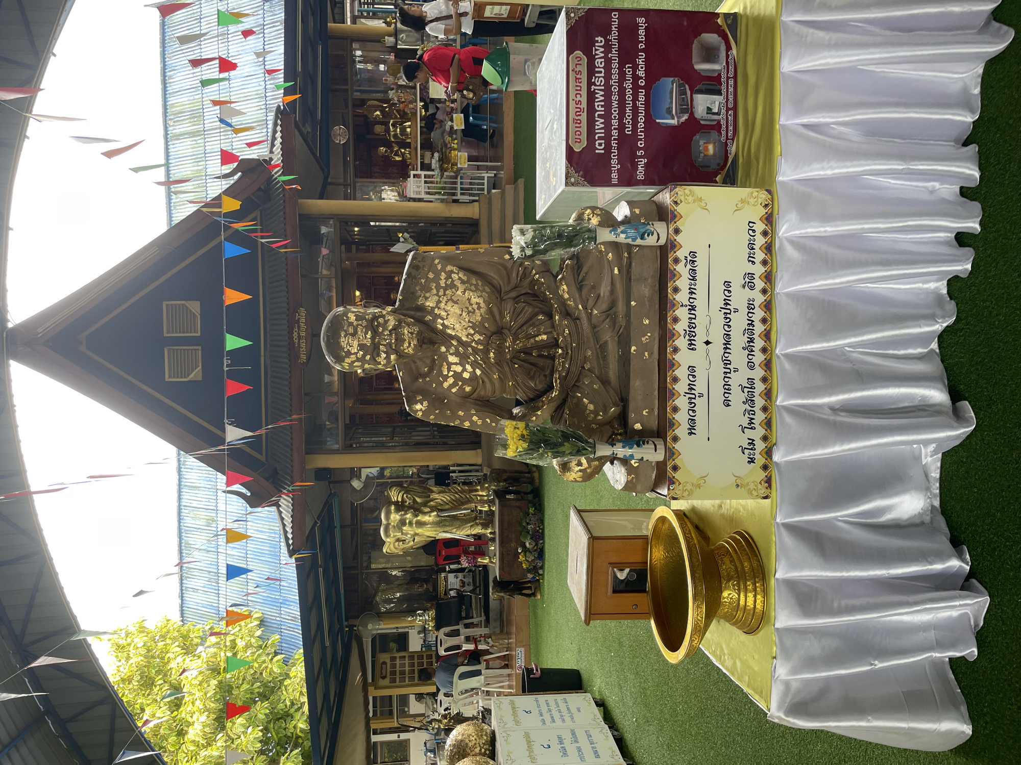 The image depicts a scene at a temple or shrine in Bang Sare, Thailand. In the foreground, a large, gold-leaf-covered statue of a monk is centrally positioned. In front of the statue, a golden bowl rests on a table draped with a white, satin-like tablecloth. Behind the statue, a box with a slot on top and a small compartment is visible, alongside a wooden structure that might serve as an offering box. The ground is covered in artificial green turf. In the background, a golden elephant statue stands prominently, flanked by smaller elephant figures. The architecture features a multi-tiered roof with colorful triangular flags strung overhead. Several people are present in the background, some appearing to be visitors or temple staff. The overall environment suggests a religious or cultural gathering place within the town of Bang Sare.