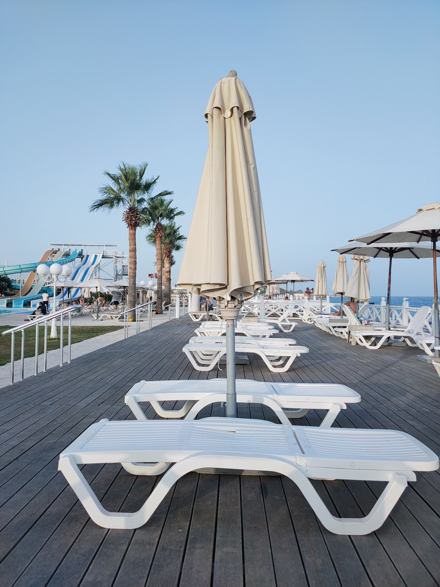 An outdoor shot captures a tranquil scene on a wooden deck, likely at a resort in Nabeul, Tunisia, under a clear blue sky. In the foreground, rows of empty white plastic lounge chairs are neatly arranged. A tall, closed beige umbrella stands prominently in the center, its fabric covering shielding it from the sun. Beyond the chairs, a white railing lines the edge of the deck, overlooking the blue sea. Palm trees with lush green fronds are spaced along the embankment. In the distance, a water park with colorful slides is visible, suggesting a family-friendly atmosphere. A few people can be seen lounging on chairs or walking near the water park, but the overall impression is one of quiet serenity, perhaps in the late afternoon or early morning.