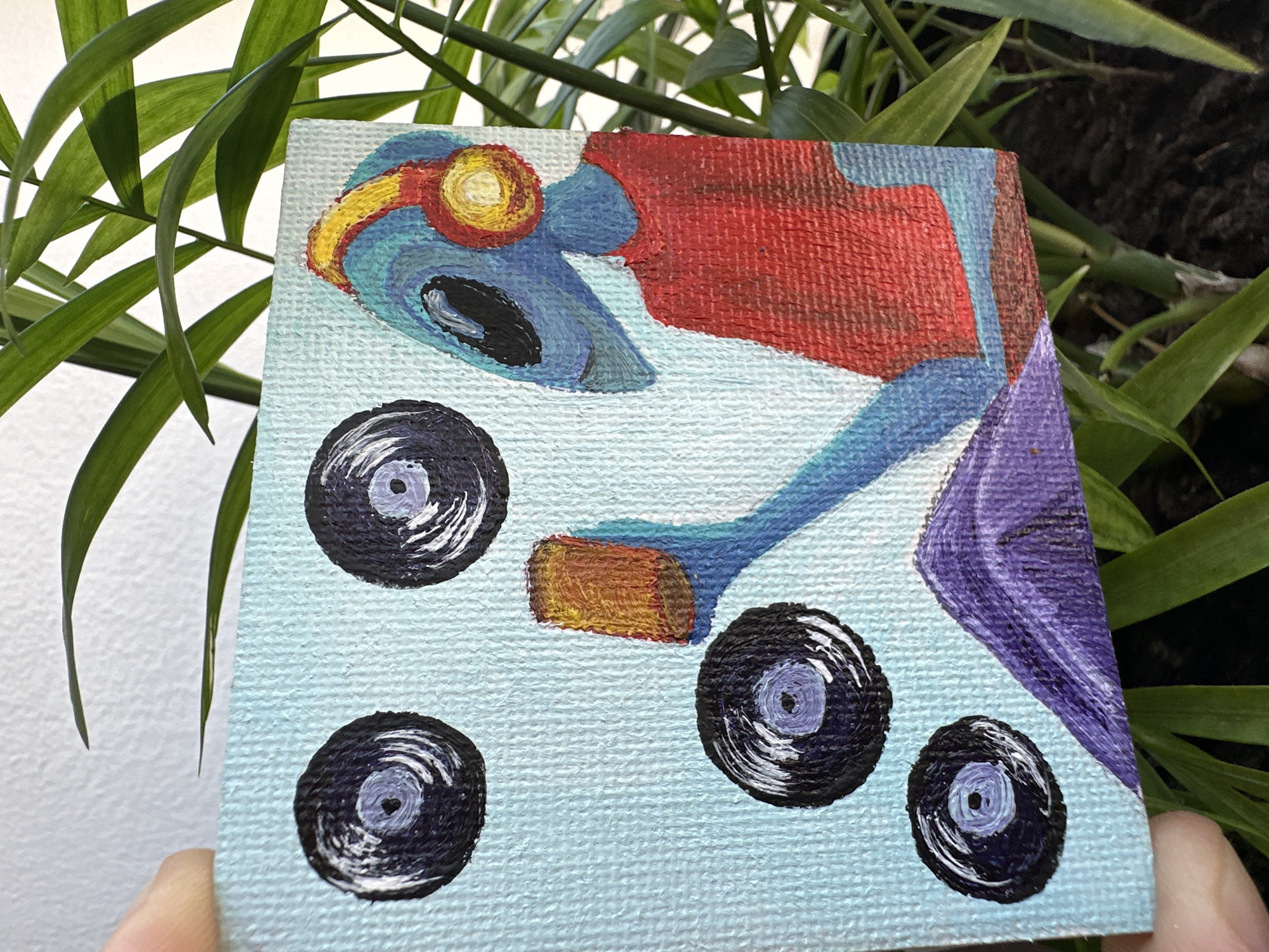 This is a close-up, eye-level, outdoor shot of a small, hand-painted canvas depicting a blue alien wearing yellow and red headphones and a red shirt, sitting and holding a tan cylinder. Four black vinyl records with purple centers are depicted on a light blue background. The canvas is positioned among lush green foliage, obscuring most of the background. The scene is not located in Dagomys, Russia.