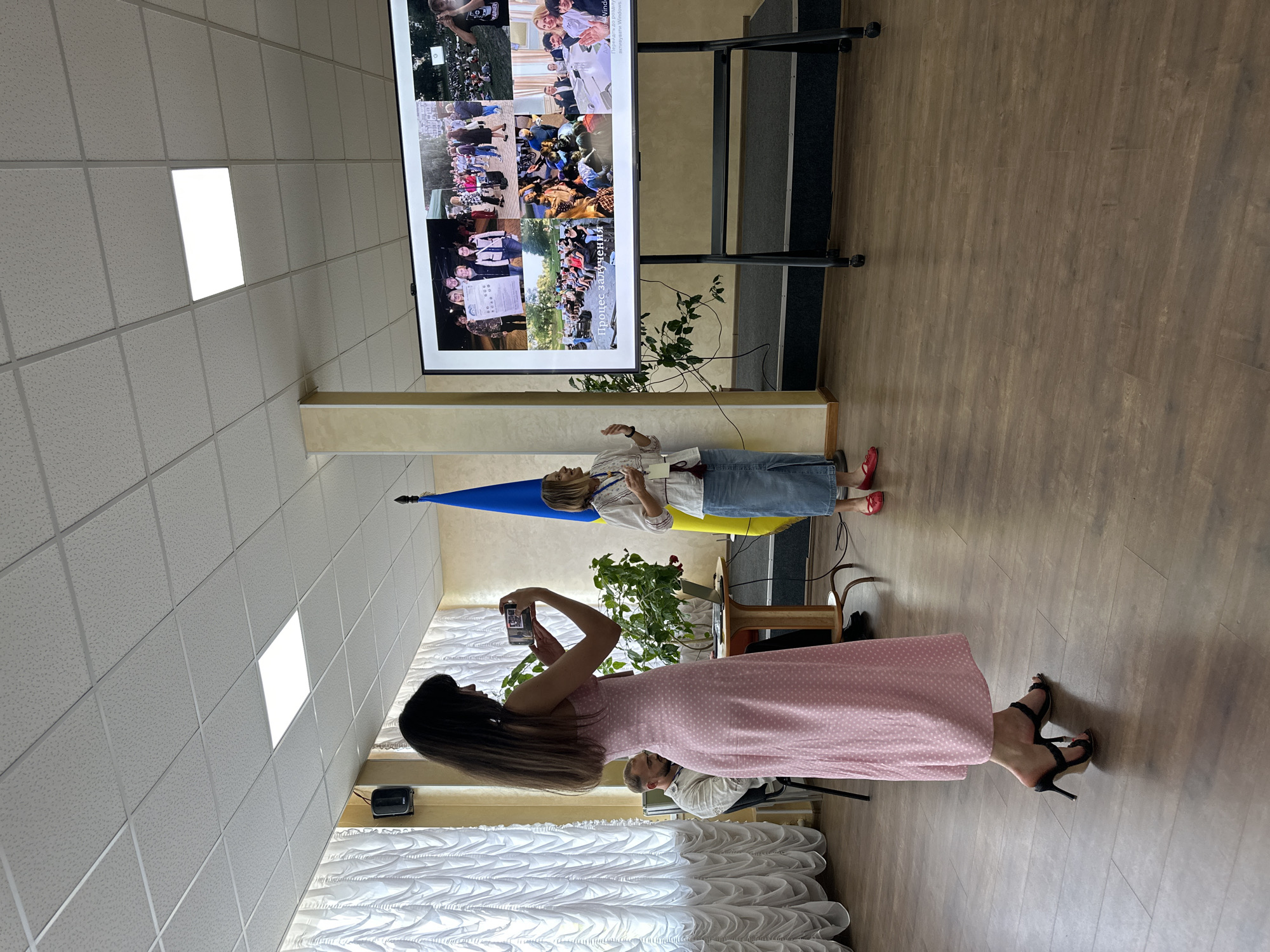 The image depicts an indoor scene in Myronivka, Ukraine, likely a presentation or gathering. Two women are the central subjects. One woman, wearing a pink polka-dot dress and black heels, stands in the foreground, holding a smartphone to take a photo. She has long, dark hair. The second woman, standing behind a Ukrainian flag, wears a white embroidered shirt and a denim skirt, holding a piece of paper. She appears to be speaking or presenting.
In the background, a large screen displays a collage of photographs, featuring many people in various outdoor settings, some holding signs. The setting includes a wooden floor, a drop ceiling with fluorescent lighting, and a plant. The overall mood seems to be one of an organized event or educational session. The presence of the Ukrainian flag suggests a connection to Ukrainian culture or current events. There is no visible indication of the time of day or weather conditions. One of the cropped images shows some text on the screen, which appears to be Ukrainian: "Процес залучення".