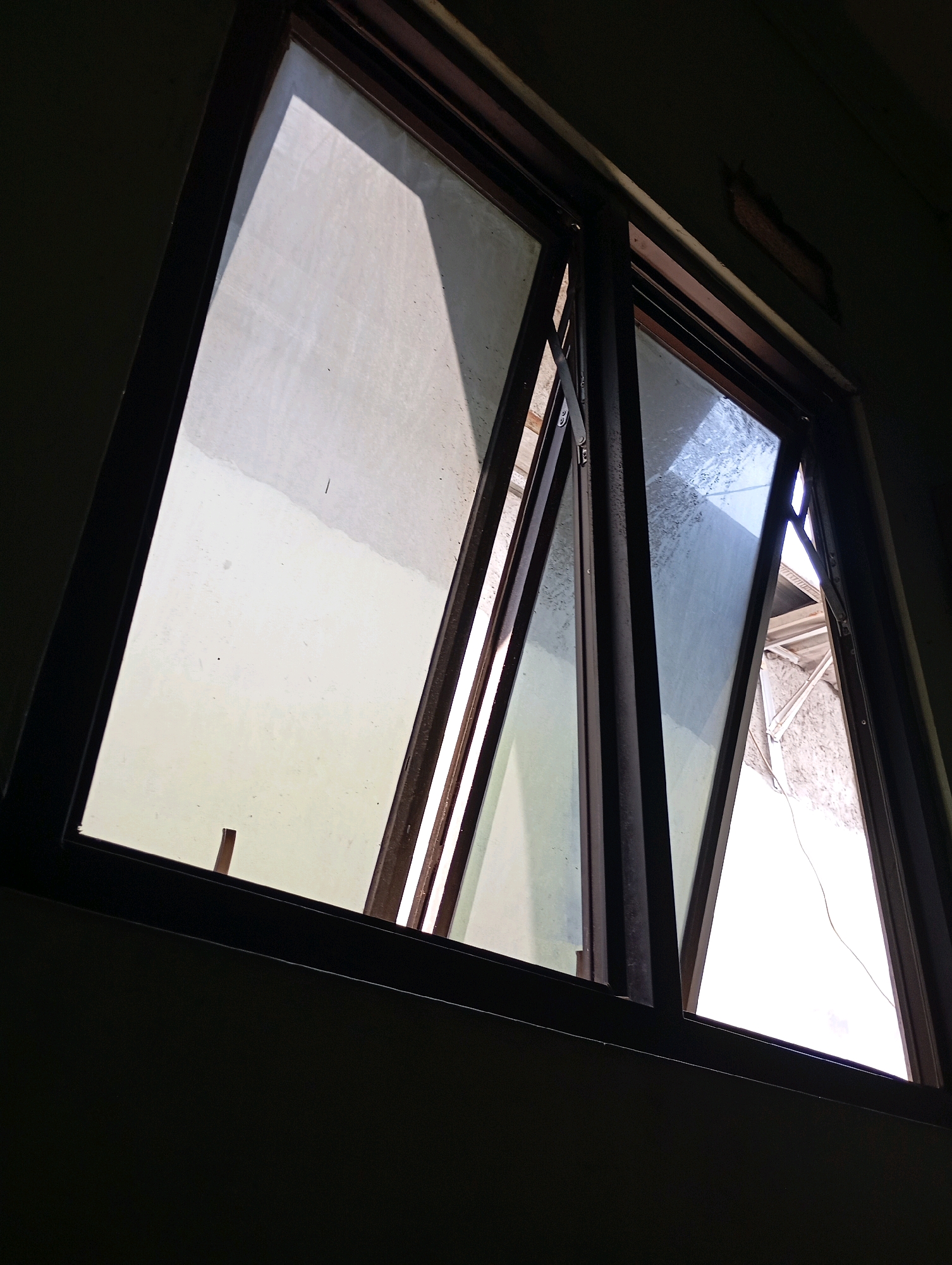 The image shows an interior view of a window in Babelan, Indonesia. The window is open, with two panes angled outwards. The glass in the panes is somewhat dirty and smudged. Sunlight streams through the opening, casting shadows on the interior wall. The window frame is dark brown. Outside, a textured wall and some roofing materials are visible. The overall scene is dimly lit, suggesting it might be daytime but the room is not directly illuminated.