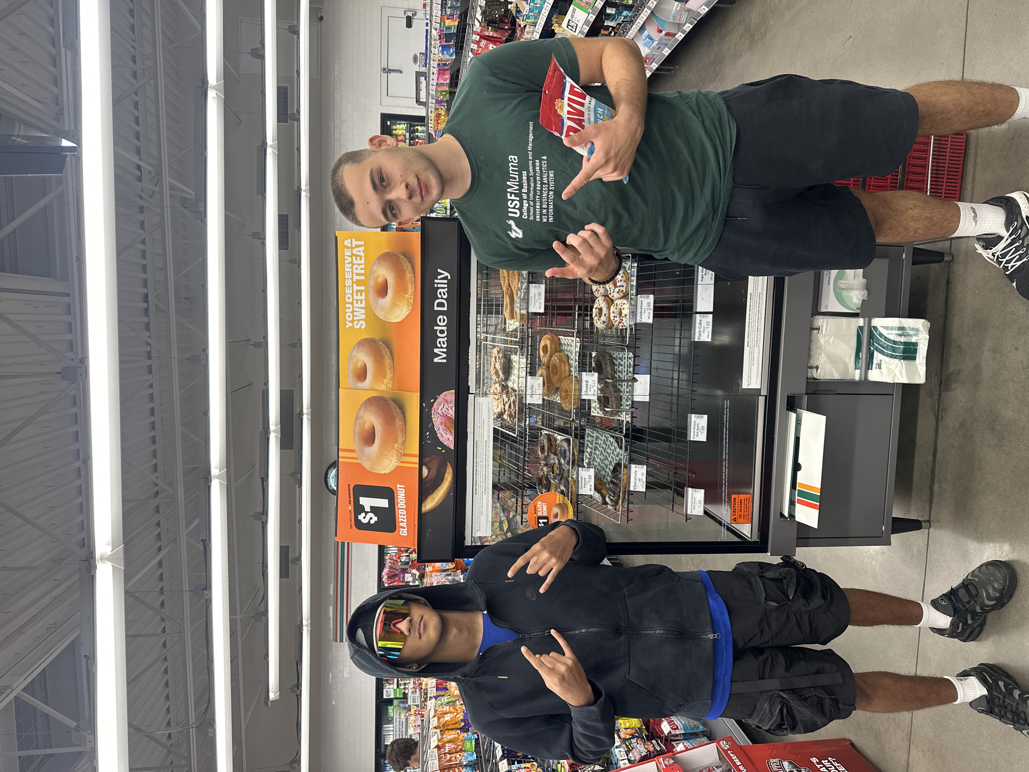 This media file depicts two young men posing inside a brightly lit convenience store, likely a 7-Eleven, in Homosassa Springs, United States of America. The image is oriented sideways, but if viewed upright, shows them standing in front of a donut display.
The man on the right, wearing a dark green t-shirt and black shorts, holds a red bag of "DAVID" brand peanuts and makes a "rock on" hand gesture with both hands. His shirt prominently displays "USFMuma College of Business School of Information Systems and Management UNIVERSITY of SOUTH FLORIDA MS IN BUSINESS ANALYTICS & INFORMATION SYSTEMS". He is also wearing white socks and black and white athletic shoes.
The man on the left, clad in a black hooded sweatshirt, black shorts, and mirrored sunglasses, also makes a similar hand gesture. A blue shirt is visible under his hoodie, and he wears white socks and black athletic shoes.
Behind them is a tiered display stand showcasing various donuts, including "GLAZED DONUT" for $1, as well as Apple, Celebration, Cream Cheese, and Plain Cake donuts priced between $1.69 and $1.99. A large yellow sign next to the display reads "YOU DESERVE A SWEET TREAT" and "Made Daily". Shelves filled with other packaged goods and drinks are visible in the background, typical of a retail store setting. Fluorescent lights illuminate the ceiling. Other visible text includes "CAUTION TIP HAZARD" on the display, and "WHAT'S YOUR BEEF?" on a red sign at the bottom. The men appear to be casually posing for a photo.