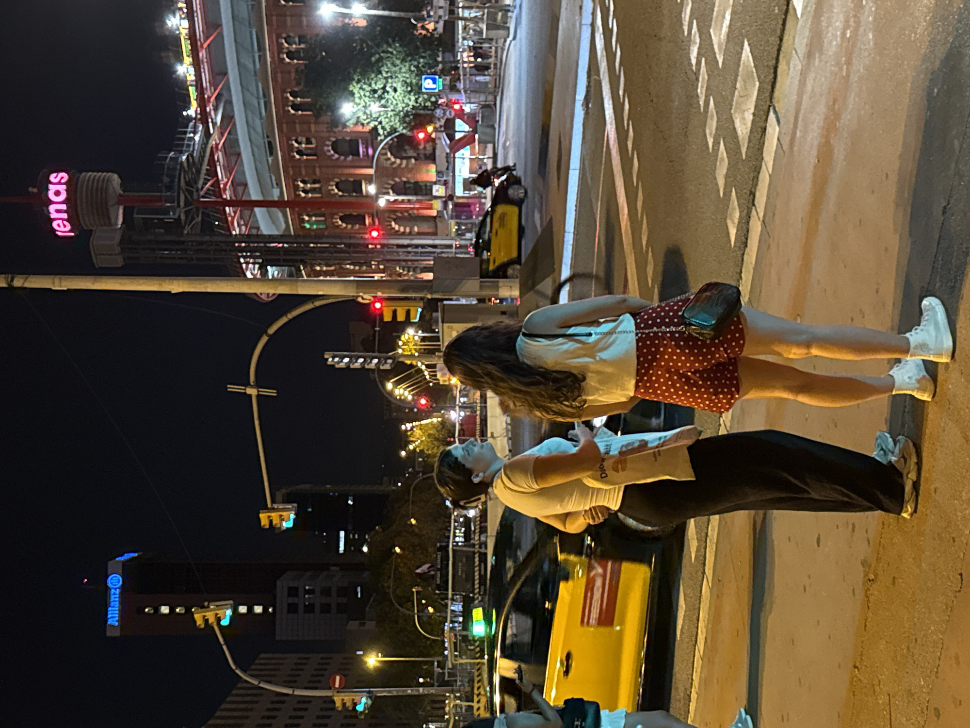 The image captures a nighttime scene on a street in Barcelona, Spain, near the Arenas de Barcelona shopping center. Two women stand on a pedestrian crossing, facing away from the camera. The woman on the left wears a white top and black pants, holding a white tote bag. The woman on the right is dressed in a white top and red polka dot shorts, with a dark purse hanging from her shoulder. A yellow taxi is visible in the background, its green taxi light illuminated. The Arenas de Barcelona, a former bullring now a shopping mall, is lit up in the background, with the word "Arenas" visible in pink neon lights at the top of its distinctive tower. Traffic lights and streetlights illuminate the urban environment. The general atmosphere is that of a busy city at night.