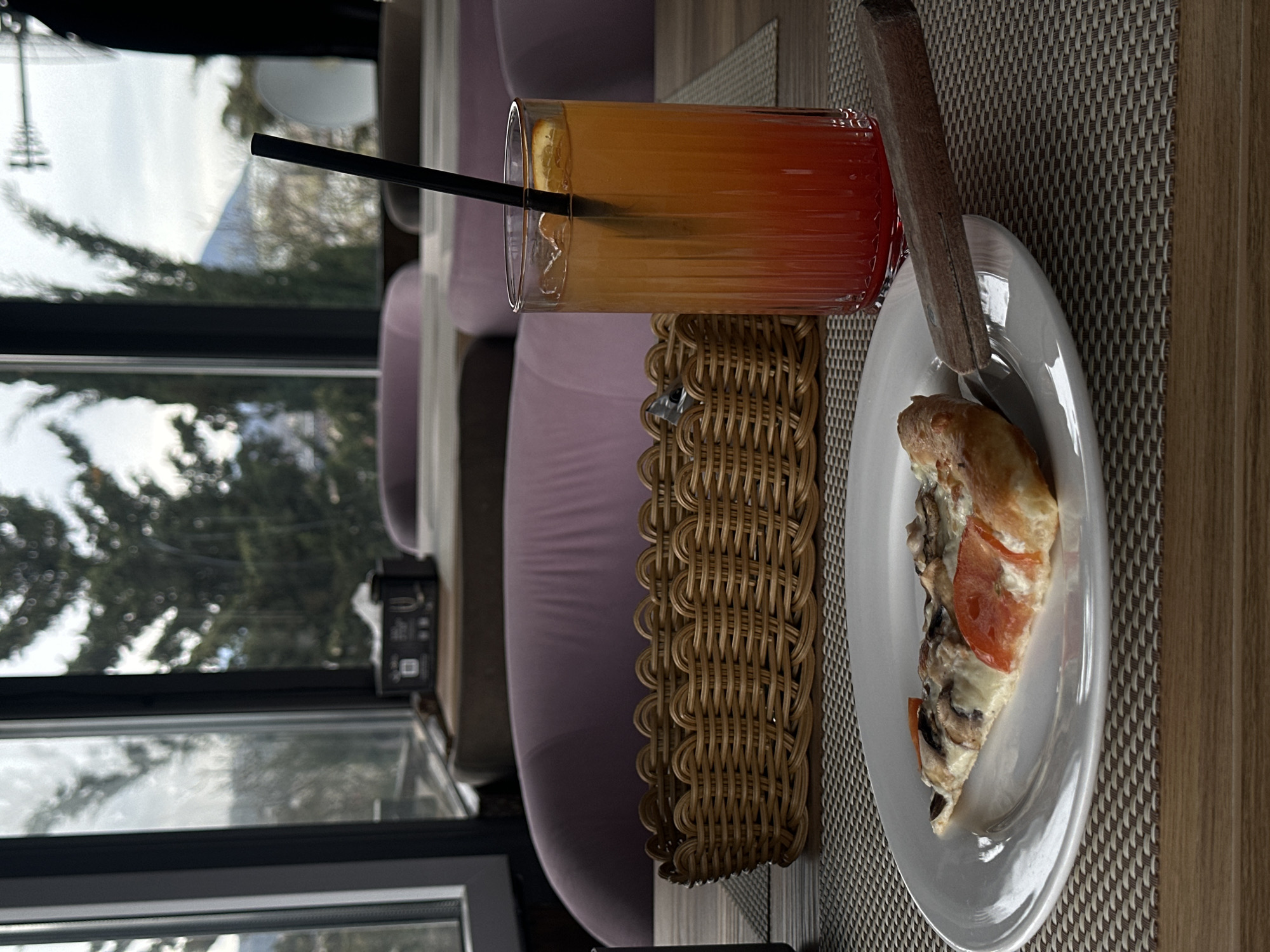 A glass of layered orange and red liquid with a black straw stands next to a small, round white plate with a slice of pizza, set on a woven placemat, with a wooden utensil between them. The setting appears to be a restaurant or cafe interior, visible through a window showcasing trees and possibly a mountain in the distance. The image appears to be taken indoors near a window. The location might be Sochi, Russia, as indicated by the prompt.