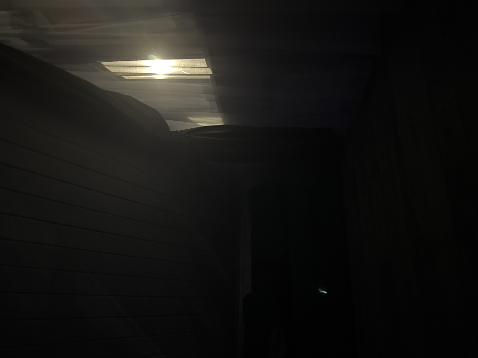 The image shows an interior scene with dark lighting. A bright light source is visible through sheer white curtains covering a window on the right side of the frame. The ceiling, on the left, appears to be made of wooden planks. The floor is dark and reflective. In the lower left corner of the image, a small, faint green light is visible, possibly an indicator light on an electronic device. The scene suggests an indoor environment, potentially a room, in Doma 1096 Km, Russia, at a time when external light is entering through a window. No individuals or specific actions are depicted.