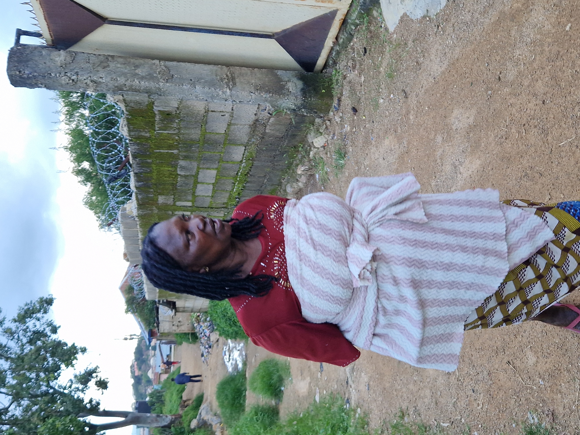 A woman with dreadlocks, wearing a red shirt and a patterned wrap, stands on a dirt road. She is looking to her right. Behind her, a concrete wall with barbed wire runs along the top. A metal gate is visible to the right of the wall. The background shows a hilly landscape with buildings and trees, suggesting an urban or semi-urban setting in Jos, Nigeria. The overall scene appears to be outdoors during the daytime, with a cloudy sky.
