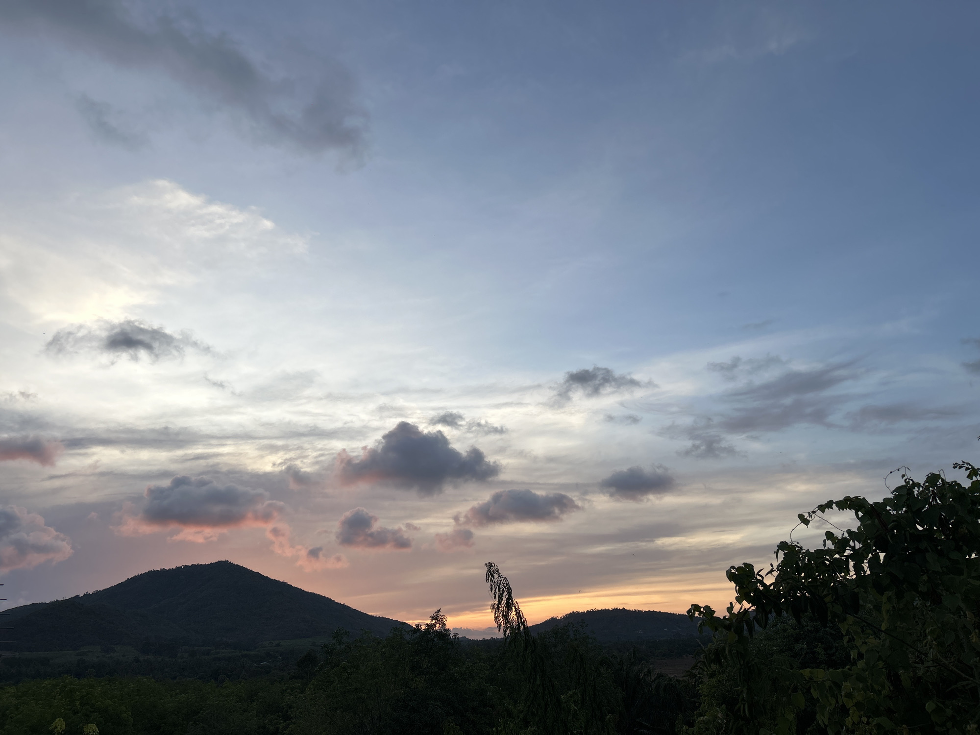 The image depicts a twilight scene near Baan Harn, Thailand. In the foreground are trees and shrubbery. In the middle ground, there are gently rolling hills. The sky is partially cloudy, with hints of orange and pink at the horizon, suggesting sunset. The upper part of the sky is a mix of light blue and grey.