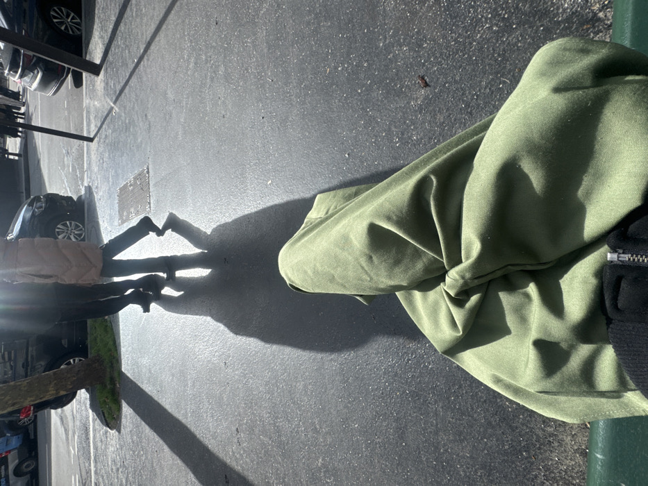 An urban scene in Paris, France, is depicted from a first-person, seated perspective. The immediate foreground reveals the photographer's legs, draped in olive-green fabric, with a black garment and a silver zipper visible at the bottom edge. A dark green surface, likely a bench, is partially visible beneath the fabric. The midground consists of a dark gray paved surface, possibly a sidewalk or street, reflecting bright sunlight. Long, distinct shadows are cast across this pavement. Two individuals are observed walking from left to right across the paved area. The individual on the left wears dark trousers and dark shoes. The individual on the right wears a light-colored, padded overcoat, dark trousers, and dark shoes. Their elongated shadows are prominent on the ground. To the left of the paved area, a small tree with a green base is present. A rectangular metal grate is embedded in the pavement to the right of the walking figures. In the background, multiple vehicles, including a dark SUV and a partially visible blue and white vehicle, are parked along a street. The scene is brightly illuminated by direct sunlight, producing a lens flare in the upper left quadrant.