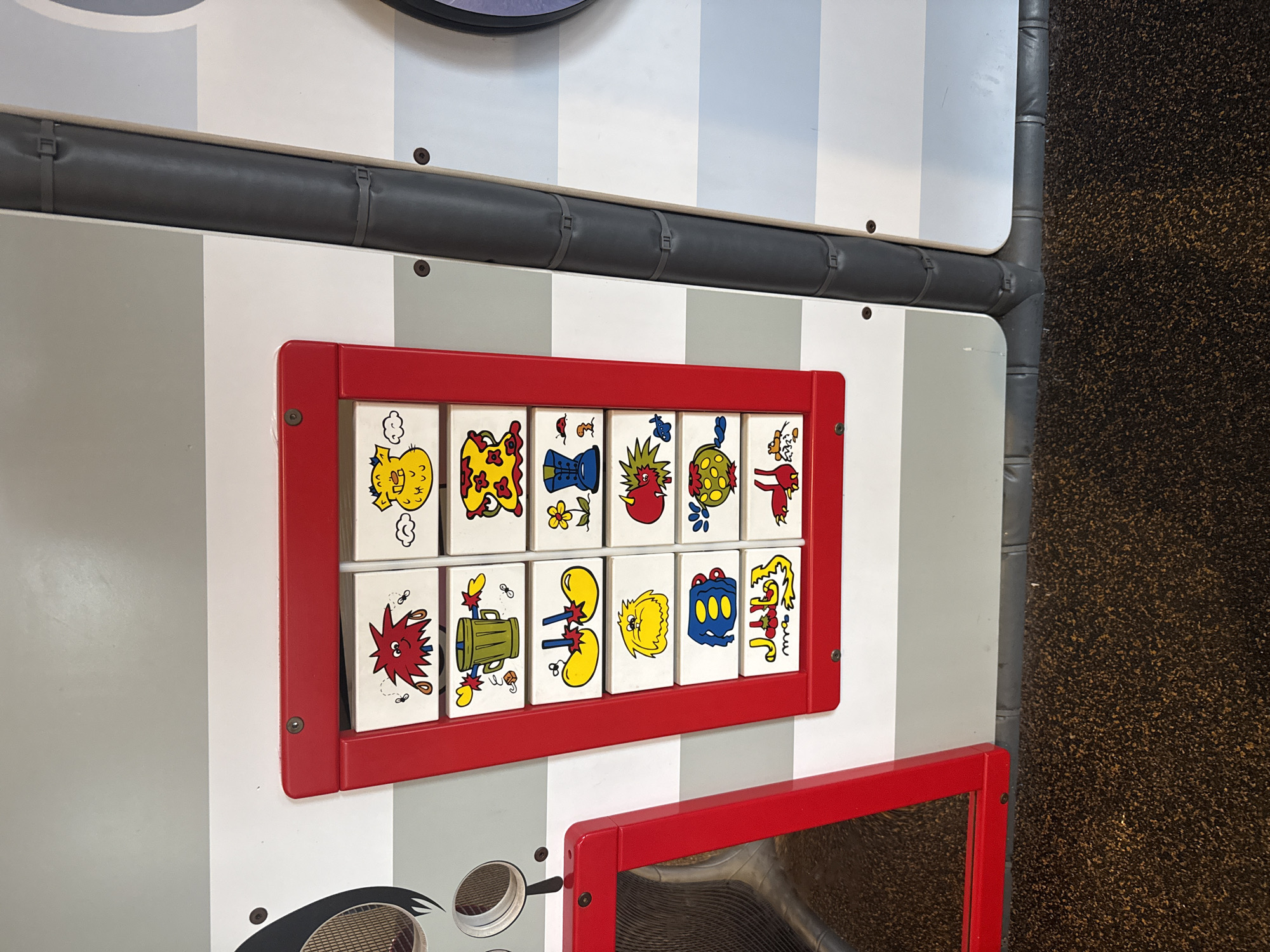 The image shows a detail of a children's play area, likely indoors, with a striped white and gray wall. A red frame holds twelve square tiles, each displaying a cartoon-like image, arranged in two rows. Below this, a partially visible red frame surrounds what appears to be a mirror or another play element. A padded, gray, cylindrical safety barrier runs horizontally above the red-framed tiles. The floor on the right is textured and dark, possibly carpeted. The location is possibly Stone Mountain, United States of America. There are no people visible in the image. The focus is on the play equipment and its details.