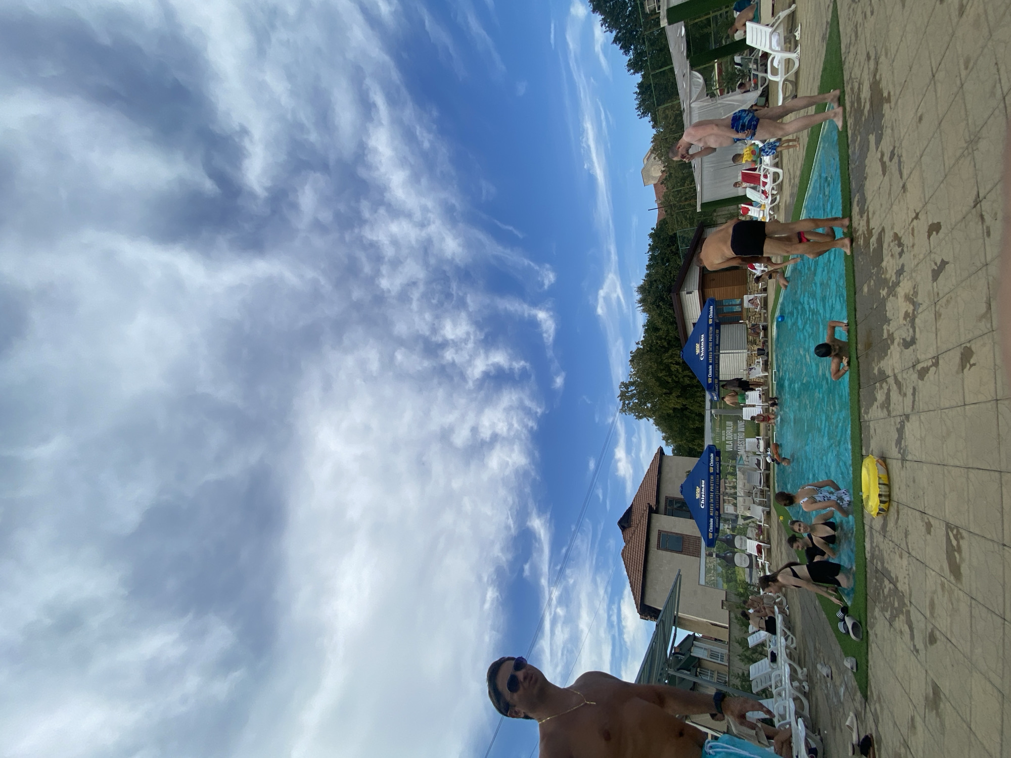 The image depicts a scene at a swimming pool located in Molovata Nouă, Moldova. In the foreground, a man with sunglasses and a gold chain stands shirtless, looking towards the camera. The swimming pool is filled with people, including children and adults, engaged in various activities such as swimming and socializing. Some individuals are standing at the edge of the pool, while others are partially submerged in the water. Several lounge chairs are arranged around the pool, some occupied by people. In the background, there are buildings with tiled roofs, umbrellas providing shade, and lush green trees. The sky above is a clear blue with scattered white clouds, indicating pleasant weather. The overall atmosphere appears relaxed and recreational, characteristic of a summer day at a pool.