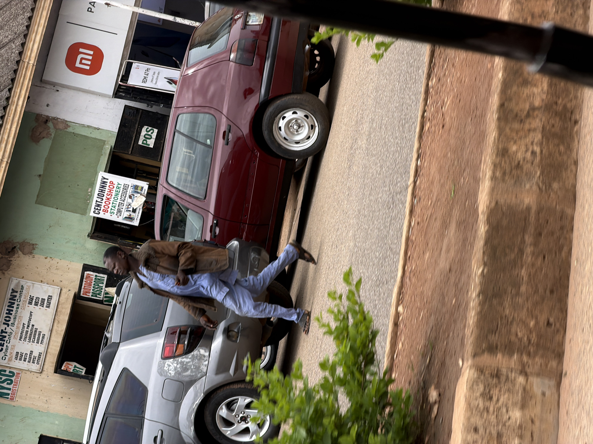 The image shows a man in a brown jacket and light blue pants walking past two cars parked on a street. The man is wearing sandals and a bag is slung over his shoulder. The cars are a burgundy minivan and a silver SUV. In the background, there are buildings with signs. One sign is for "Xiaomi" with a red logo. Another sign says "POS". A sign for "CENT-JOHNNY BOOKSHOP • STATIONERY • COMPUTER ACCESSORIES" is visible. There is also a sign that says "PHOTOCOPY PASSPORT". The scene appears to be in an urban environment, possibly a commercial area, given the signs for businesses. The lighting suggests it is daytime. The overall impression is a snapshot of daily life in a Nigerian street scene.