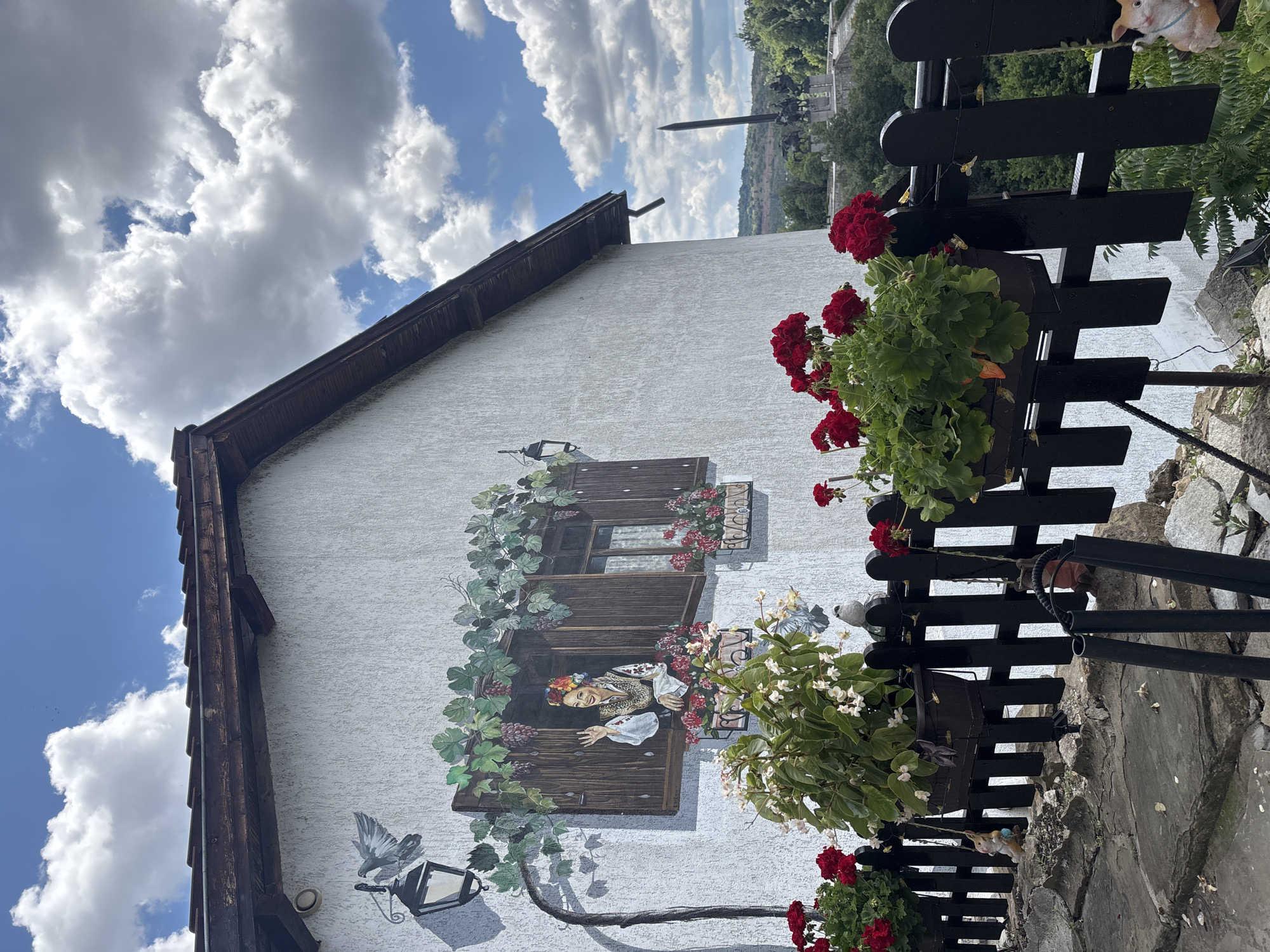 The image depicts a close-up view of a building's exterior wall in Veliko Tarnovo, Bulgaria, featuring a mural. The mural shows a woman in traditional Bulgarian attire waving from an open window, surrounded by painted grapevines and potted flowers. Red geraniums are blooming in actual planters along a black wooden fence in the foreground. In the background, a monument with a tall obelisk and equestrian statues is visible, situated on a hill overlooking a green, wooded landscape under a partly cloudy sky. The building has a white, textured wall and a dark, sloped roof. The scene is likely a residential or commercial property in the historic city.