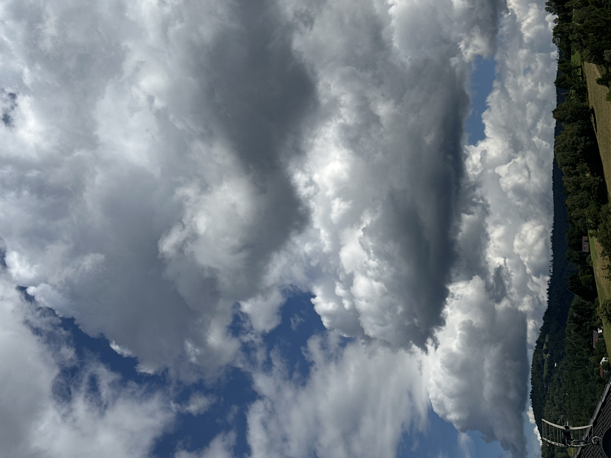 The image captures a view of a cloudy sky above a hilly, forested landscape. The sky is filled with a mix of white and dark grey cumulus clouds, with patches of blue sky visible. In the foreground, a rooftop with a large satellite dish is visible on the left. The middle ground features rolling hills covered in dense green trees, interspersed with some scattered houses and fields. The horizon line is dominated by the dark silhouette of the tree line against the sky. The overall impression is of a rural or suburban setting under a dramatic sky. The location is Wisła, Poland.