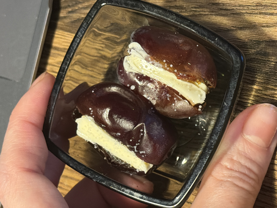 A light-skinned hand grips a small, square, transparent dish with dark edges. Inside the dish, two dark brown dates are presented. Each date has been longitudinally cut open and filled with a creamy, off-white substance. White, granular crystals are sprinkled over the dates and their filling. The dish is positioned over a wooden surface displaying a distinct grain pattern. A portion of a grey electronic device is visible in the upper right background. The scene is located in Perm, Russia.