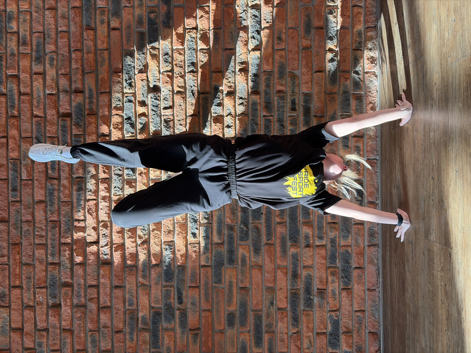 An individual is depicted performing a handstand indoors against a textured brick wall in Chaikovsky, Russia. The person, appearing female with light-colored hair, is positioned centrally in the frame. They are wearing a black short-sleeved t-shirt with yellow text, which reads "SPORT FEST" upside down, black sweatpants with elasticated cuffs, white socks, and white athletic shoes. One leg is extended vertically towards the top of the frame, while the other leg is bent at the knee, with the foot near the mid-thigh of the extended leg. Both hands are placed flat on a light brown wooden floor with visible plank lines, supporting the body. Strong, directional sunlight enters from the top right, casting sharp shadows on both the brick wall and the wooden floor, highlighting the textures of the surfaces and the contours of the individual's clothing. The brick wall features a pattern of reddish-orange and darker grey bricks.