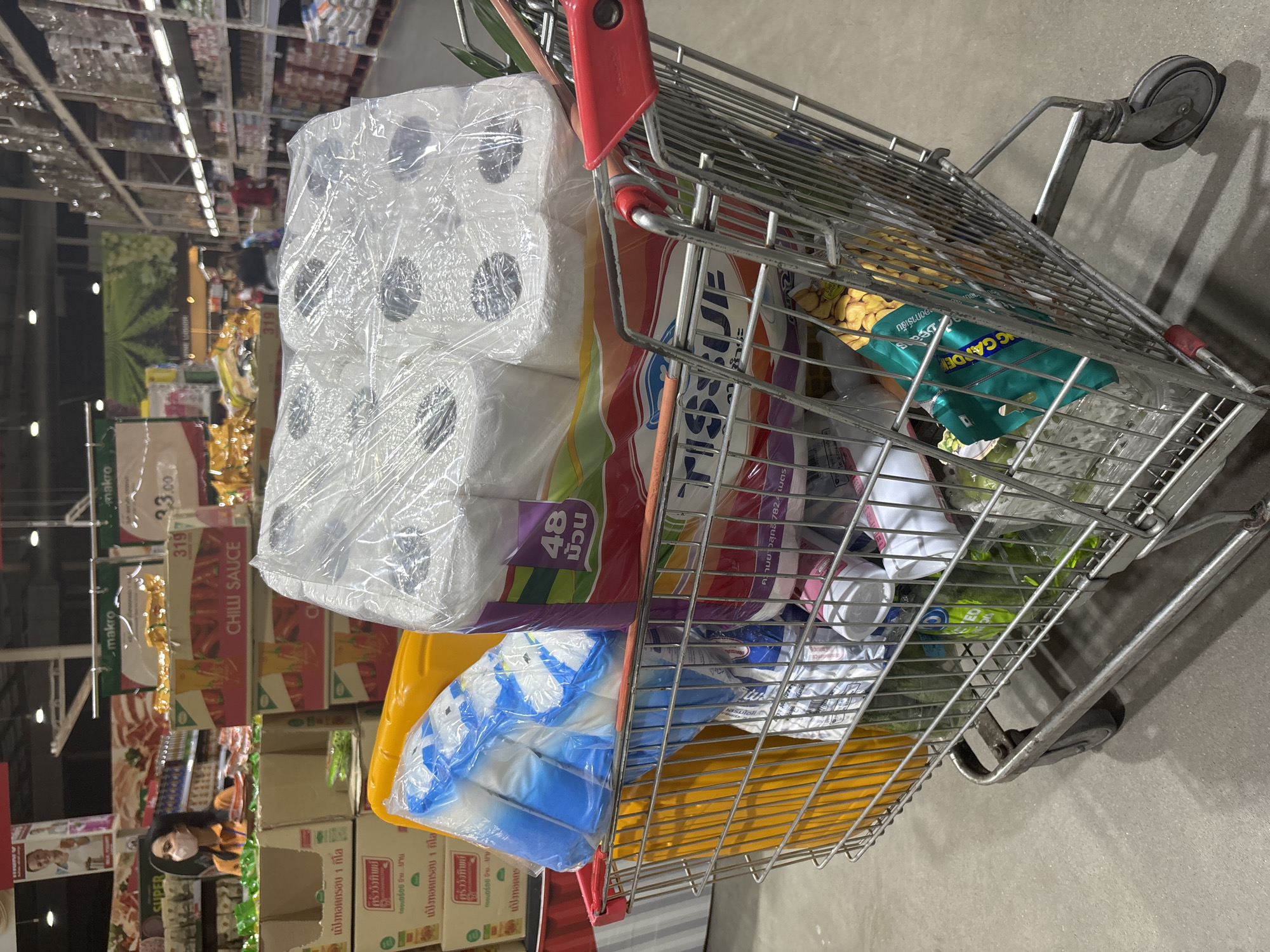 The image shows a shopping cart in what appears to be a grocery store or supermarket in Baan Bo Phut, Thailand. The cart is filled with various items, including a large package of toilet paper (labeled "48 ม้วน" which translates to "48 rolls"), a package of TISSUE, and other packaged groceries. Shelves lined with more products are visible in the background, along with a person wearing a face mask. Signage including prices ("33.00") and product names ("CHILLI SAUCE") in both English and Thai are visible on the shelves.