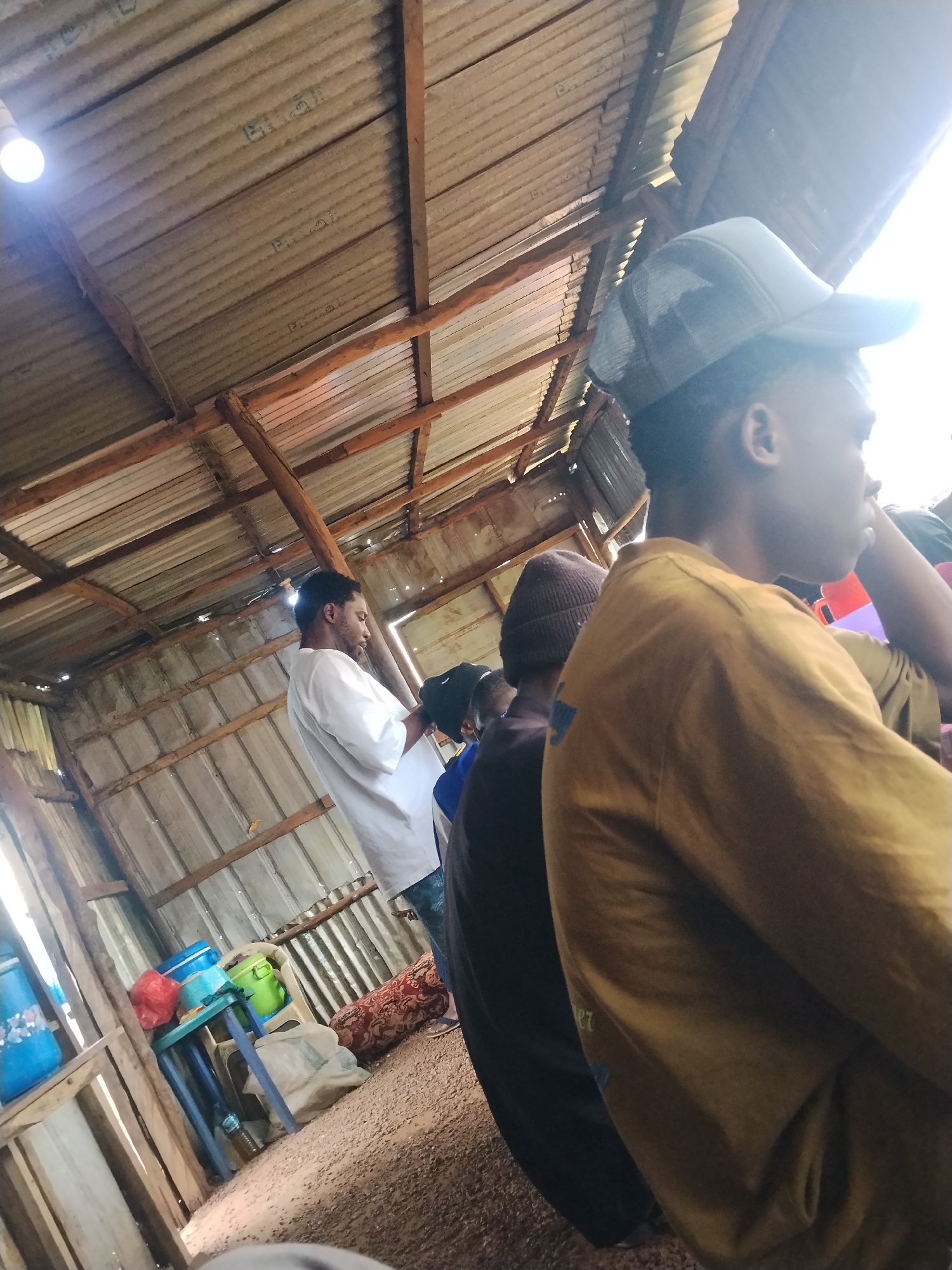 The image depicts an interior scene, possibly a market stall or a workshop, in Durumi, Nigeria. The structure has a corrugated metal roof supported by wooden beams. Sunlight streams in from the right, illuminating dust motes and creating a bright, hazy atmosphere. Several individuals are present. In the foreground, a person wearing a mustard-colored t-shirt and a grey mesh baseball cap is seen in profile, their face partially obscured by the cap and bright light. To their left, another person in a dark t-shirt and a brown beanie hat is partially visible. Further in the background, a man in a white t-shirt is standing and looking downwards. To the left of the frame, there are various objects including blue and green plastic containers, a red plastic bag, and a patterned bolster cushion. The floor appears to be made of packed earth or sand. The overall impression is one of a casual, informal gathering in a rustic setting.