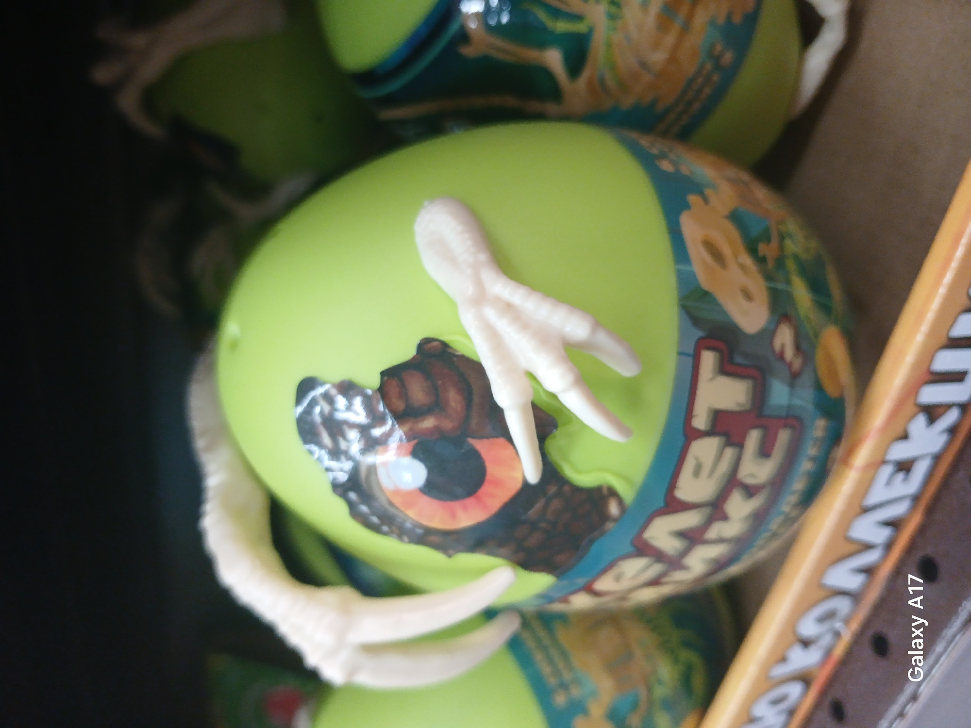 The image shows a close-up view of several green plastic eggs, likely toys, in a container or box. The egg in the foreground has a printed design on it featuring a dinosaur eye breaking through a shell. Protruding from the egg are molded white plastic claws. The brand name "Dino Egg" is visible. The lighting suggests an indoor retail environment. In the lower right corner, the text "Galaxy A17" is visible, indicating the device used to take the photo. Although nothing explicitly shows the location, the user requested to assume the location as Yekaterinburg, Russia.