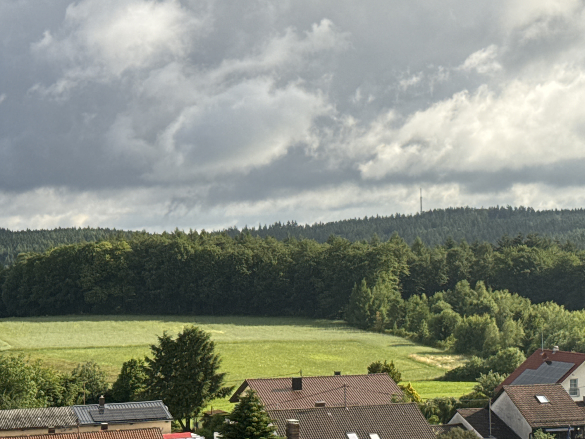 The image displays a panoramic view of a rural landscape in Strümpfelbrunn, Germany, under a dramatic, cloudy sky. In the foreground, rooftops of residential buildings with dark grey and red tiles are visible, some featuring solar panels. A tall, slender tower, likely a telecommunications mast, stands on the horizon against the cloudy sky. The middle ground is dominated by a dense, dark green forest that stretches across the frame, with a bright green field of grass or crops in front of it. The overall scene is serene and natural, with the presence of human habitation indicated by the buildings and the distant tower.
