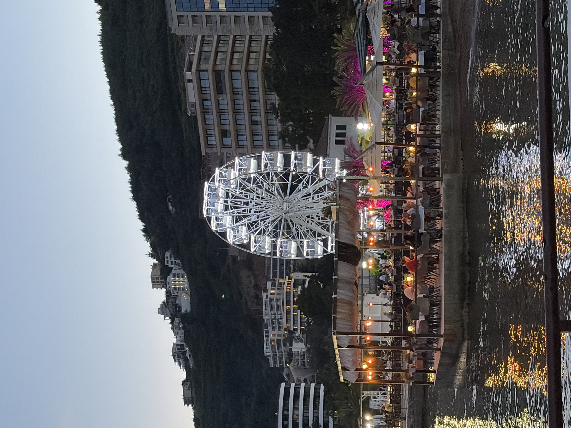 An exterior, wide shot captures a waterfront dining area in Budva, Montenegro, at dusk. A large, illuminated Ferris wheel stands prominently in the background. In the foreground, a raised platform hosts a bustling outdoor restaurant with numerous tables occupied by patrons. The tables are adorned with small lights, and umbrellas provide shade. The water in front of the dining area reflects the colorful lights from the restaurant and the Ferris wheel. The city's hilly terrain, dotted with buildings, forms the backdrop, with the sky appearing a deep blue as night approaches.