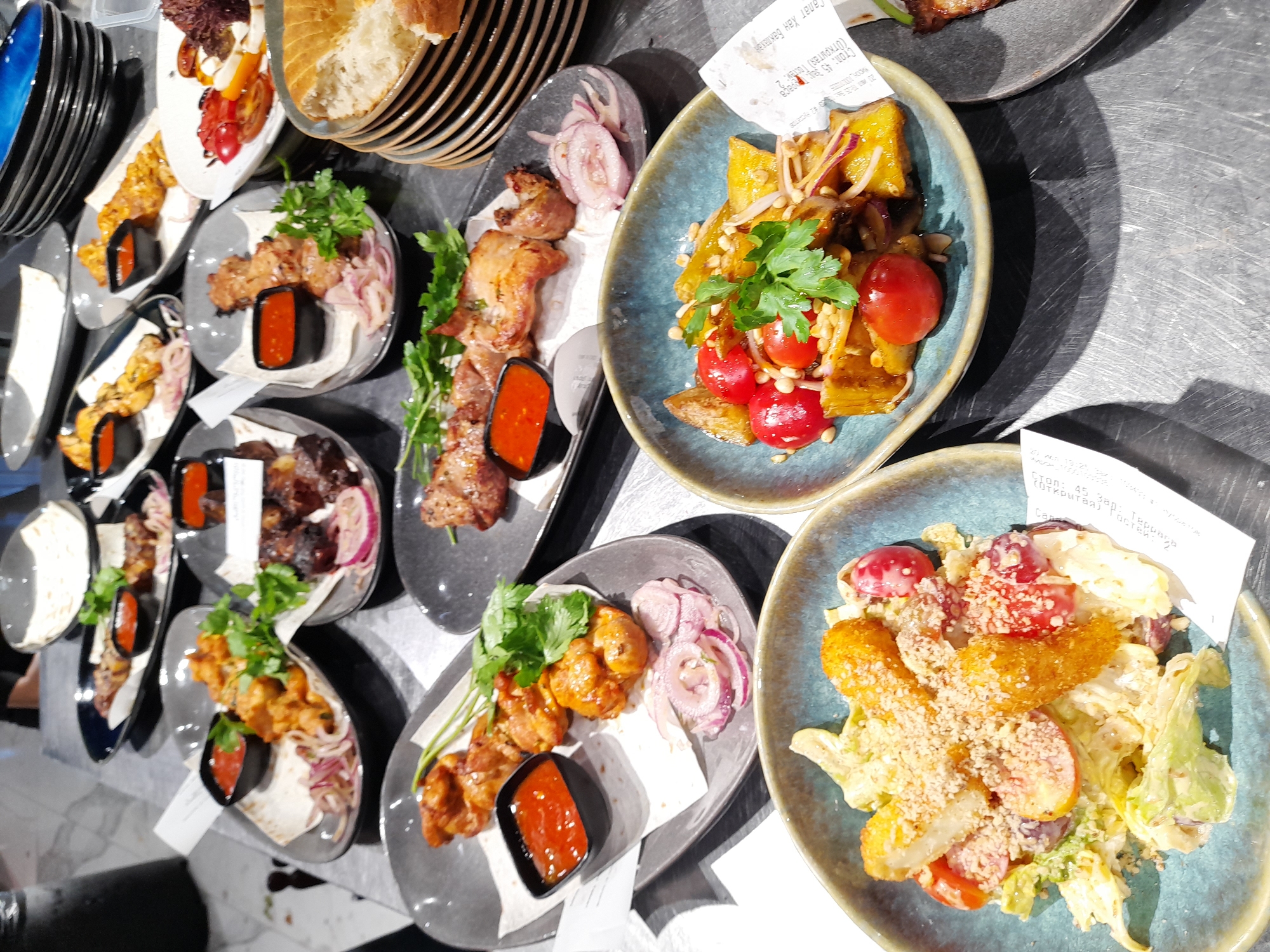 A close-up, high-angle shot displays a table laden with various dishes of food, likely at a restaurant in Vladivostok, Russia. The food includes grilled meat skewers served with flatbread and red sauce, a salad with fried pieces, and a warm vegetable dish with tomatoes and pine nuts. The table surface appears to be metal and is reflecting light, suggesting it could be an outdoor seating area or a restaurant with industrial-style decor. The overall presentation suggests a casual dining experience.