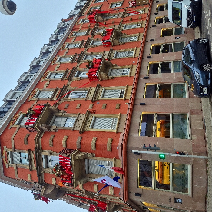 The image displays a vibrant urban street scene, primarily featuring the facades of two multi-story buildings and parked vehicles under a clear daytime sky. On the left, a prominent building with a red facade showcases numerous windows equipped with light-colored frames and external roller shutters, many of which are partially or fully closed. Ornate balconies with black wrought iron railings and beige stone supports project from several floors, festively adorned with red ribbons, greenery, and holiday decorations like stars and baubles. A security camera is visible in the upper left corner. Flags hang from the lower part of this building, including one for "Best Western" and another displaying "DA", along with red, white, and blue elements suggesting a French flag.
Adjacent to this, on the right, is a multi-story building with a lighter brown facade, identified as a "HOTEL" by a sign on its ground floor. Its large rectangular windows reflect bright sunlight or interior lighting.
Parallel to the buildings, a street features two parked vehicles: a dark blue hatchback with a partially visible French license plate (beginning with 'F') and a white van, both facing right. A green traffic light is prominently positioned in the mid-right section of the image. The decorations on the balconies suggest a holiday or festive period.