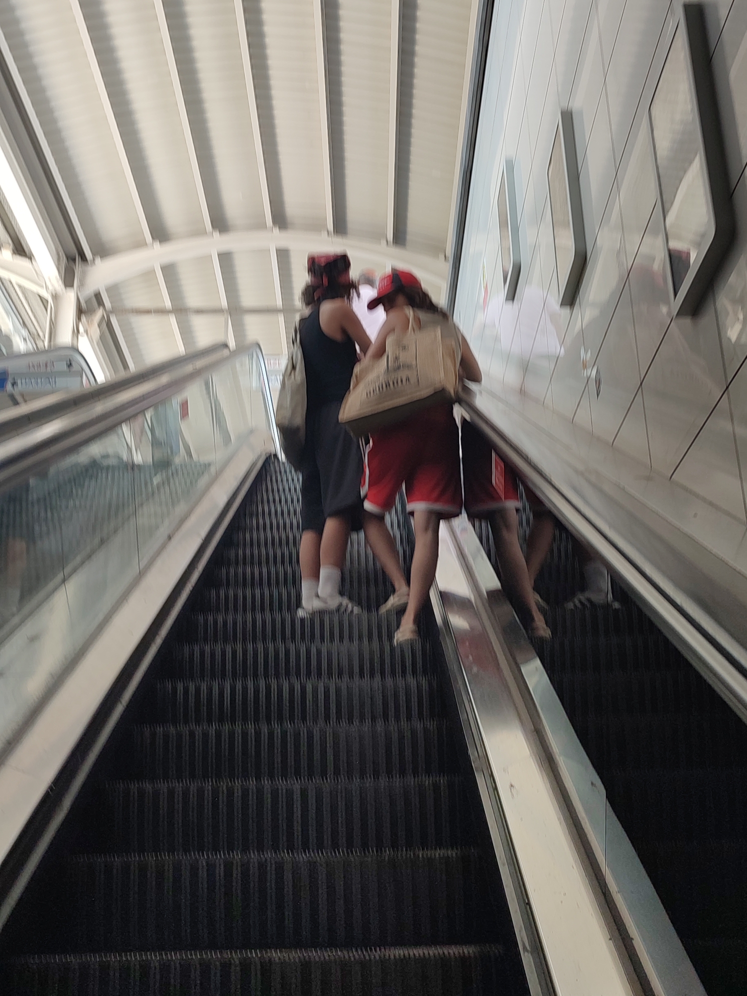 The image shows two individuals on an escalator at the Kadıköy Metro Station in Istanbul, Turkey. The person on the left is wearing a black tank top, red and black checkered headscarf, and dark shorts, carrying a light-colored bag over their shoulder. The person on the right is wearing a red cap, a white tank top, and red shorts with white trim, also carrying a light-colored bag. Both individuals are facing upwards on the escalator, which has dark gray treads and a metallic handrail. The escalator is situated under a white, ribbed ceiling structure. The wall to the right of the escalator is tiled with white square tiles, and some framed displays are visible on the wall. Reflections of the people and the escalator can be seen on the metallic surfaces.