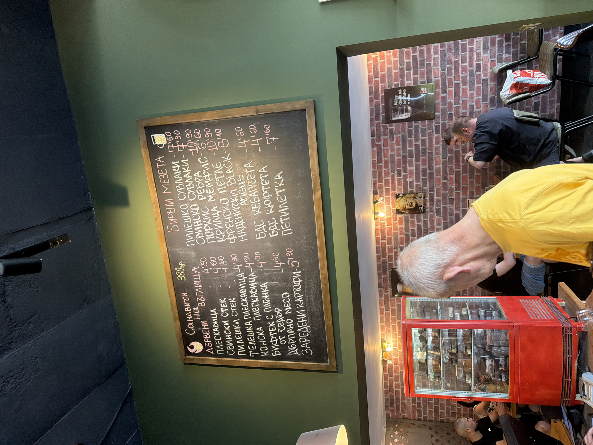 The image depicts the interior of a cafe, potentially located in Basarbovo, Bulgaria, as indicated by the text on the chalkboard menu which appears to be in Bulgarian. The focal point of the image is a chalkboard menu with handwritten items and prices. To the left of the menu, a red refrigerated display case filled with cakes is visible. In the foreground, a man with graying hair wearing a yellow t-shirt is facing away from the camera, obscuring a portion of the scene. In the background, several other patrons are seated at tables. The walls are decorated with brick-patterned wallpaper and posters. The overall ambiance suggests a casual dining establishment.
