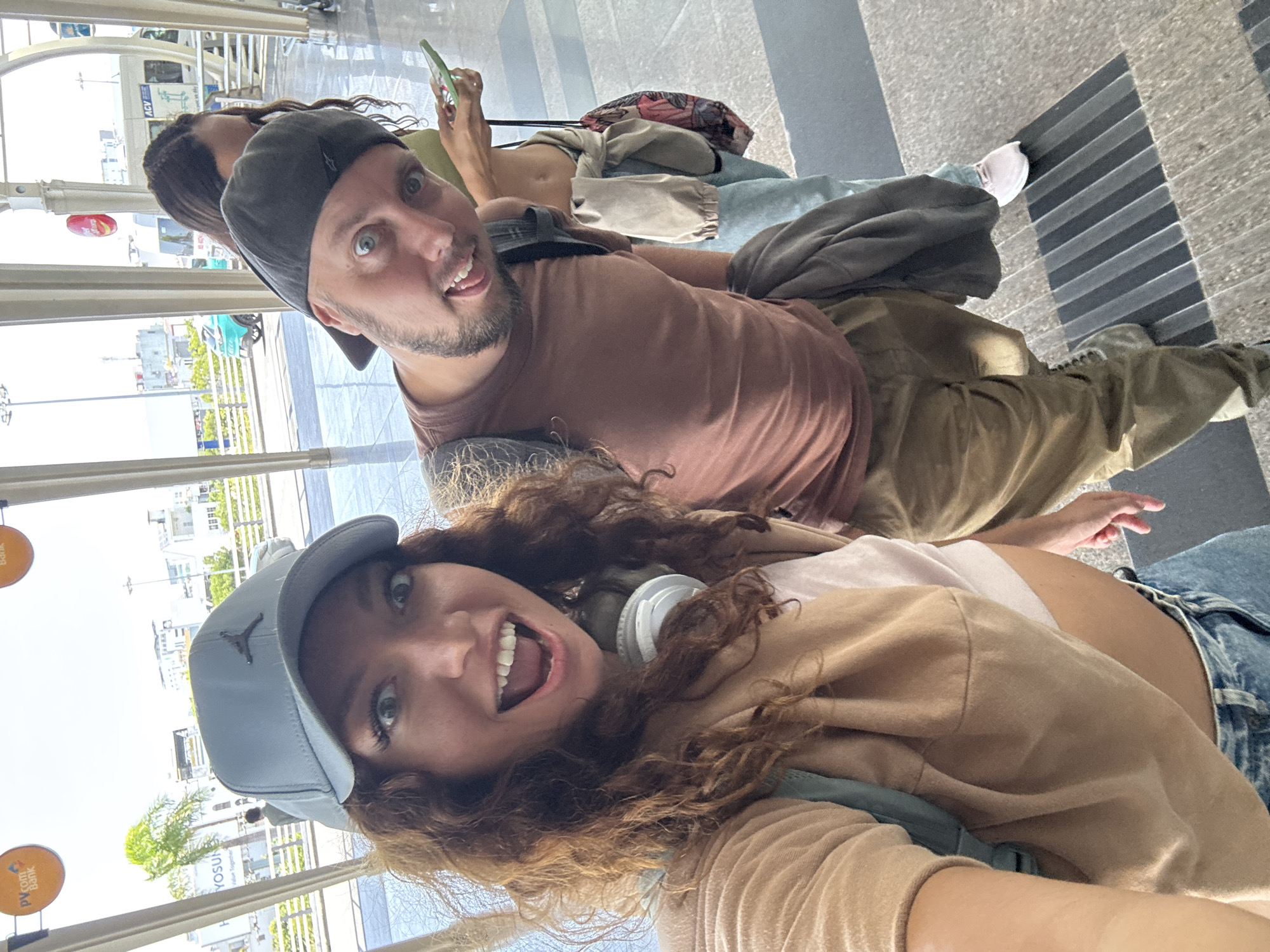 The image shows two people taking a selfie. The foreground subject is a fair-skinned female with long, curly brown hair, wearing a light blue baseball cap, a beige jacket, a white T-shirt, blue jeans, and white headphones around her neck. The background subject is a fair-skinned male with short brown hair and a beard, wearing a grey baseball cap and a brown T-shirt. Another person holding a phone sits in the background. All three people are seated and appear to be waiting. They are inside what appears to be an airport terminal with large glass windows that show a parking area, signage, and vegetation outside, and a PVComBank sign is visible. The location is identifiable as the city of Đà Nẵng, Vietnam. The foreground flooring consists of grey and black stripes.