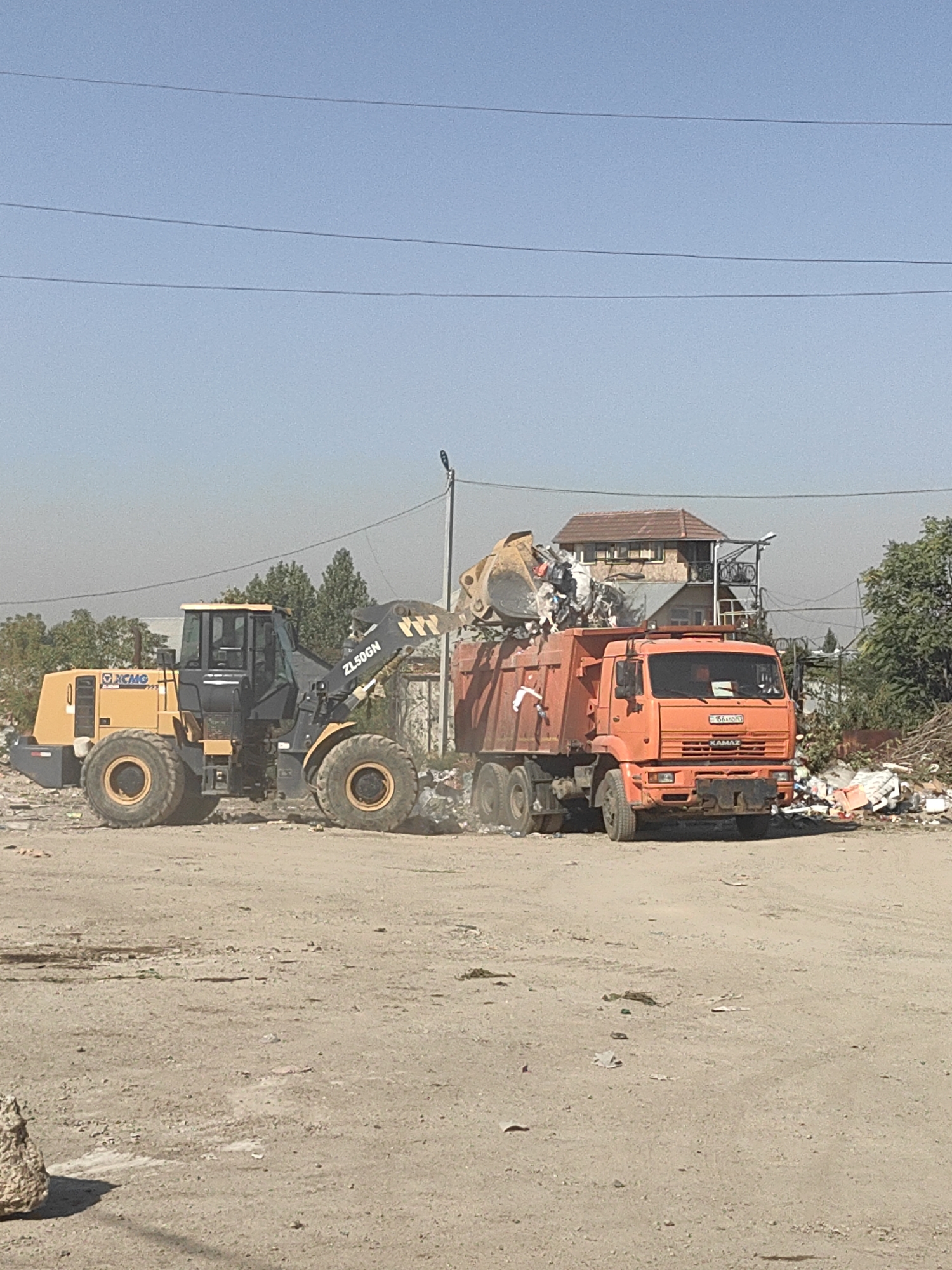 In the city of Shymkent, Kazakhstan, at a waste disposal site, a yellow wheel loader, identified by the "XCMG ZL50GN" markings, is actively loading debris into the back of an orange Kamaz garbage truck. The truck's rear is full of waste material. The environment is a dusty, open area with piles of garbage visible in the background, along with some trees and buildings. The sky is clear and blue.