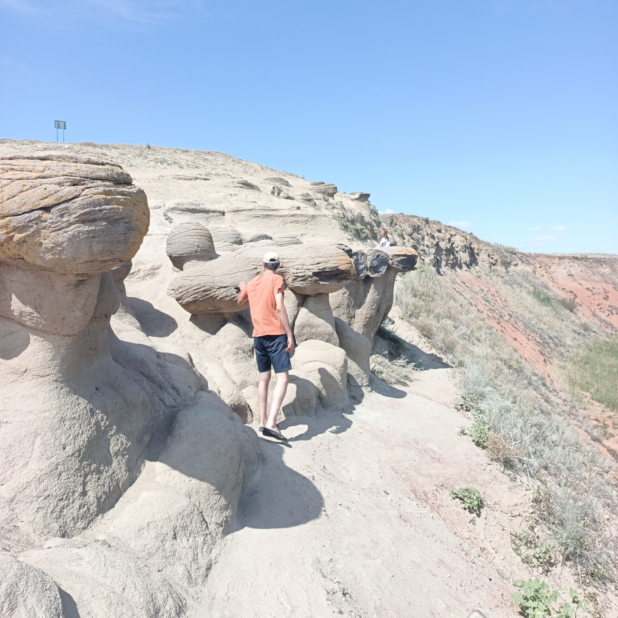 In this outdoor, daytime photograph taken in the city of Mikhailovka, Russia, a young man in an orange t-shirt and dark shorts stands on a dusty, eroded landscape composed of sandstone formations. He is facing away from the camera, looking towards a dramatic cliff edge. His right hand rests on a large, rounded sandstone boulder. The ground is dry and sandy, with sparse, dry vegetation scattered around. In the background, the landscape continues with more weathered rock formations and a steep, reddish-brown cliff face. The sky above is a clear, bright blue. Another individual, a child, is visible in the far distance sitting on a rock formation. A distant blue sign is visible on a hilltop to the left.