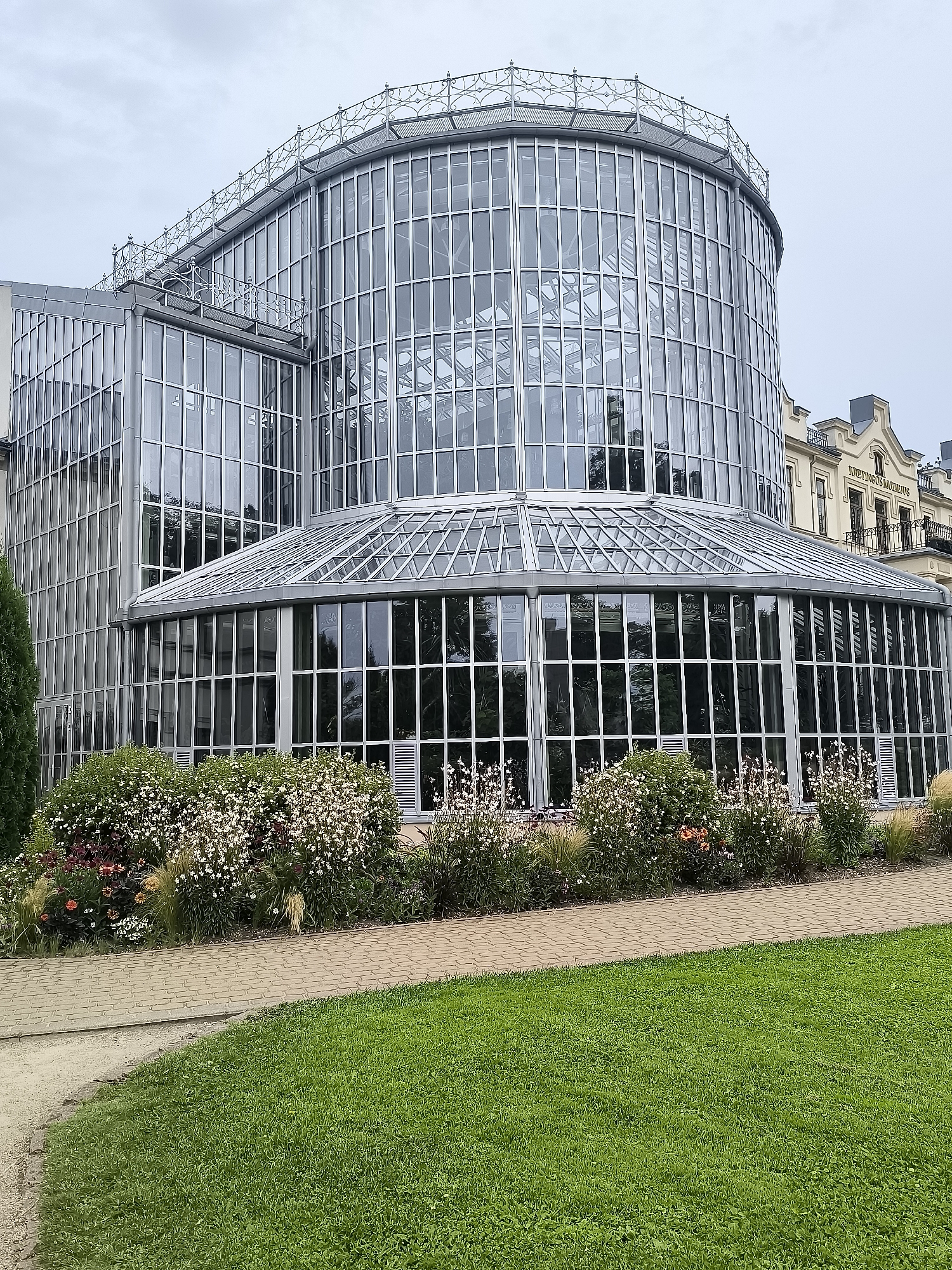 The image depicts the Kretinga Manor greenhouse, a prominent architectural feature located in Kretinga, Lithuania. The greenhouse is a large, multi-story structure constructed primarily of glass and metal, with a distinctive curved facade. It is surrounded by a manicured garden featuring lush green grass, a paved pathway, and a variety of flowering plants and shrubs. In the background, a portion of an older, ornate building with classical architectural elements is visible, suggesting the historical context of the manor grounds. The sky is overcast, casting a diffused light over the scene. There are no people visible in the image. The overall impression is one of tranquility and architectural beauty within a well-maintained natural setting.