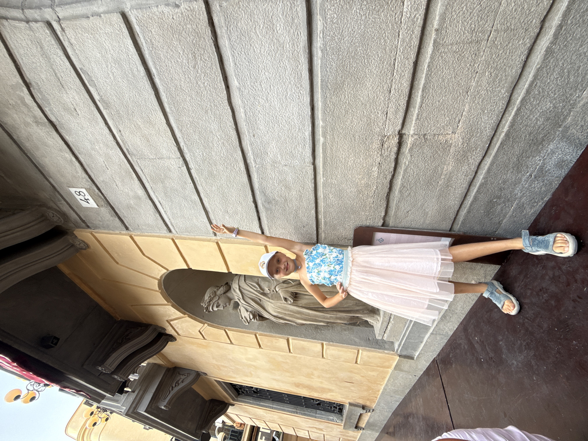 In an outdoor setting with textured grey stone walls overhead, a young girl is pictured posing. She is wearing a white cap, a blue and white floral top, a light pink pleated skirt, and blue sandals. Her left arm is extended upwards, and her right hand is on her hip. She is smiling and looking towards the camera, appearing happy and engaged.
Behind the girl, there is a niche in the wall containing a stone statue of a draped figure, possibly religious in nature. The architecture suggests a historical or cultural site. The ground in front of the girl is a dark, reddish-brown material, possibly wood or stone. To the left of the girl, architectural elements are visible, including ornate carvings and a window with a metal grate. Some yellow, cartoonish decorations, perhaps from a local festival or attraction, are also partially visible on the far left.
A small white rectangular tag with the number "48" written in black marker is attached to the stone wall above and to the left of the girl. The lighting suggests it is daytime, with bright sunlight casting shadows. The overall scene conveys a sense of tourism or leisure in a European location, potentially Mataró, Spain.