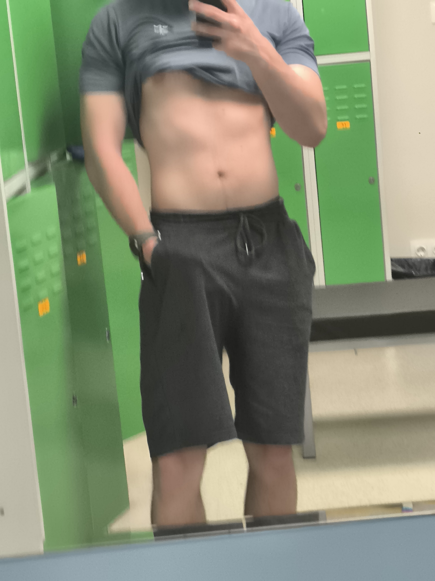 A person is standing in what appears to be a locker room, taking a selfie in the mirror. The person is wearing a grey t-shirt pulled up to expose their torso and grey sweat shorts. Their left hand is in the pocket, and a watch is visible on their left wrist. The background shows green lockers with silver handles and orange number labels. There is a bench visible in the background. The location is not possible to determine from the image provided, so I am unable to fulfill that portion of the request.