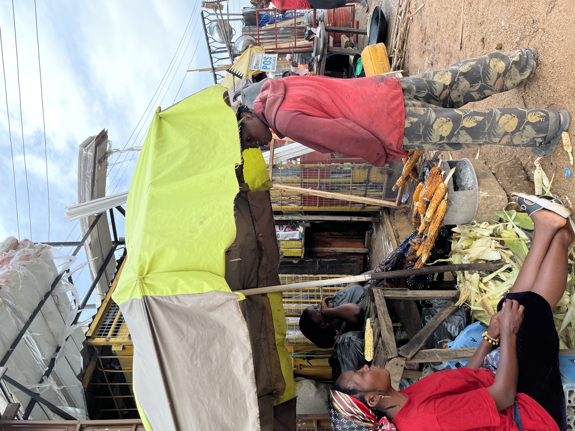 The image depicts an outdoor market scene in Gura Top, Nigeria, under an overcast sky. Two women are the primary subjects. One woman, wearing a red t-shirt and black skirt, sits on a blue plastic stool, her legs crossed, and is looking to her left. She has a colorful headscarf and bracelets on her wrist. The other woman, standing, wears a red hooded sweatshirt and patterned camouflage pants. She is facing right, looking slightly away from the camera, with her left hand resting on her hip. In the foreground, several ears of corn are being grilled on a metal stand above a charcoal brazier. There are more corn husks and ears of corn scattered around the ground. The market stall behind the women features a yellow and brown umbrella providing shade, with shelves displaying various items, including what appear to be food products in boxes. The overall environment is rustic, with a dirt ground and various makeshift structures.
